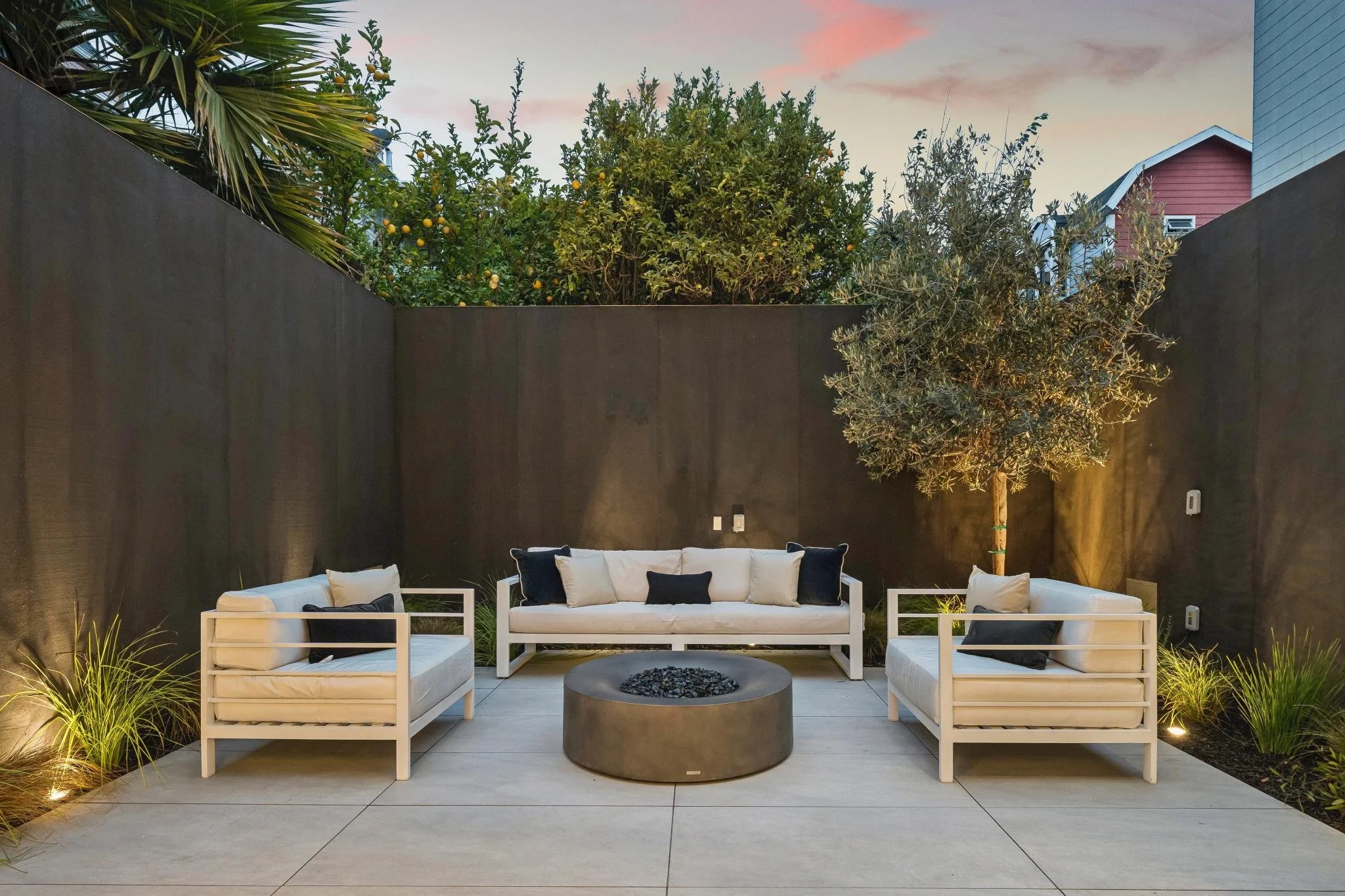 Modern backyard outdoor seating area with white cushioned furniture, black pillows, a round fire pit in the center, surrounded by landscaping, and a tall dark fence with trees and houses in the background under a pink and blue sky.