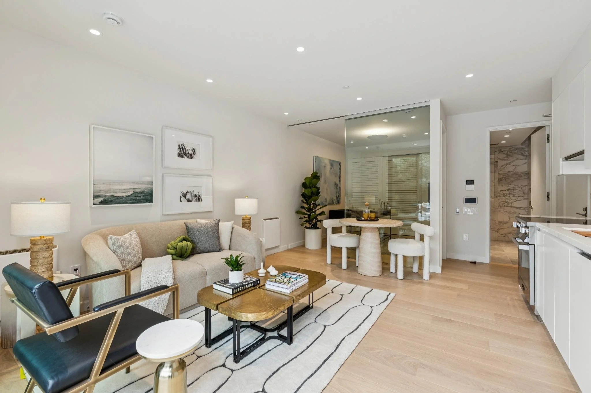 Modern living room with beige sofa, black chair, wooden coffee table, white walls, framed artwork, floor lamps, green plants, and a small dining area with chairs.