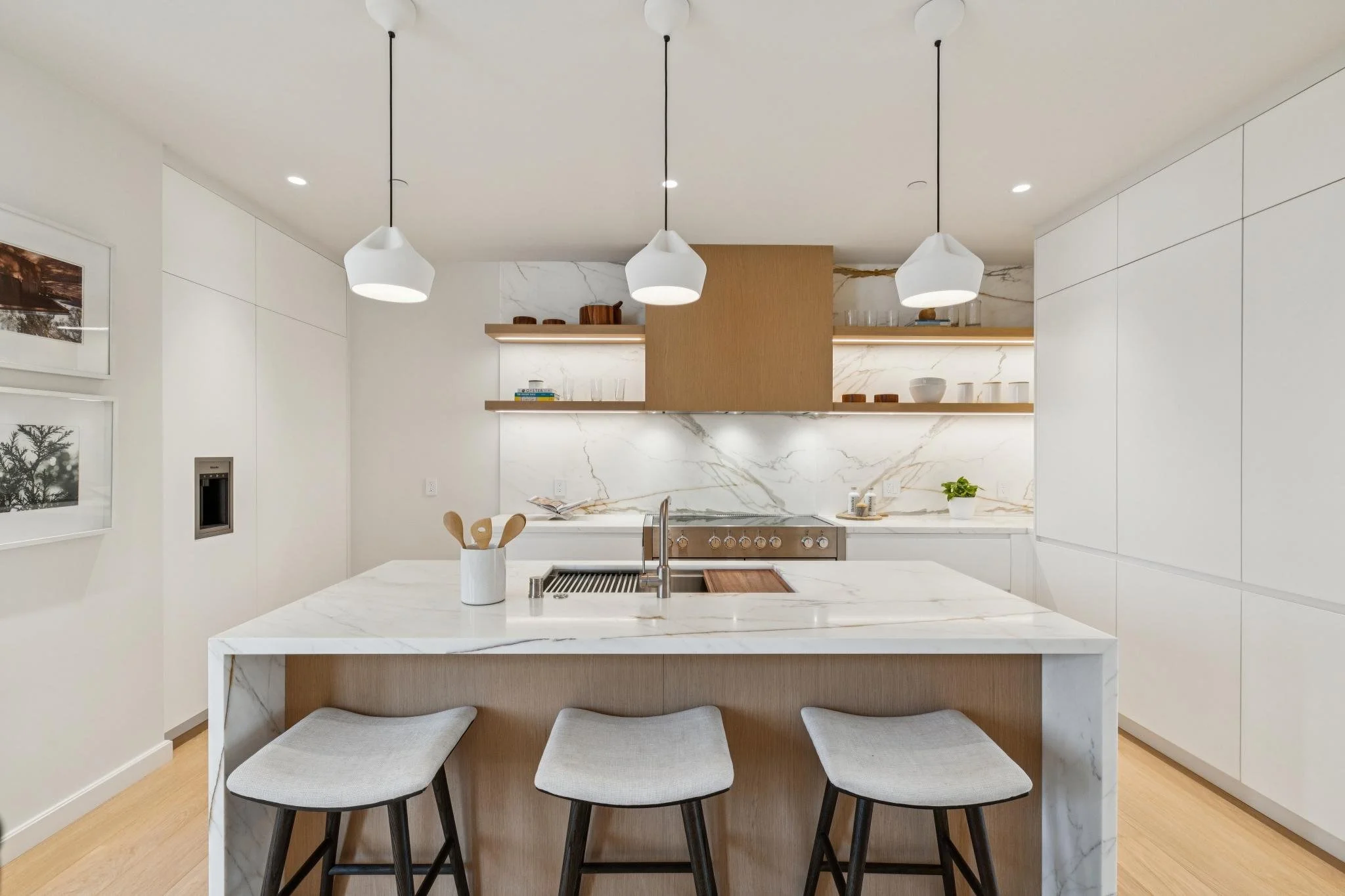 Modern kitchen with white marble island, three gray upholstered stools, white cabinetry, wooden shelves, and three hanging pendant lights.