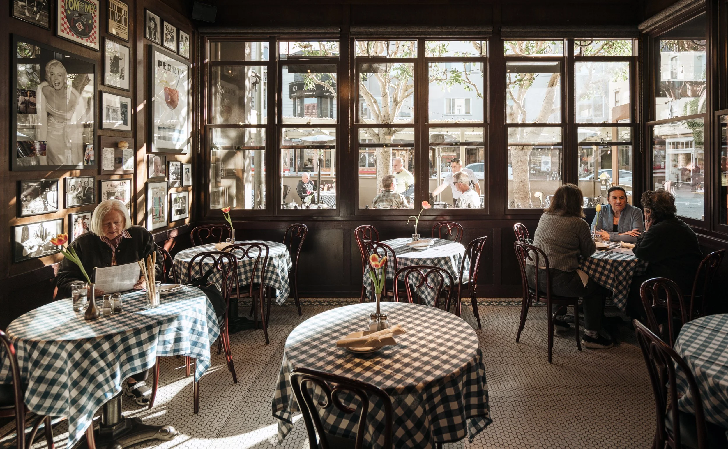 Interior of a restaurant with checkered tablecloths, flowers on tables, and large windows showing outside seating and pedestrians.