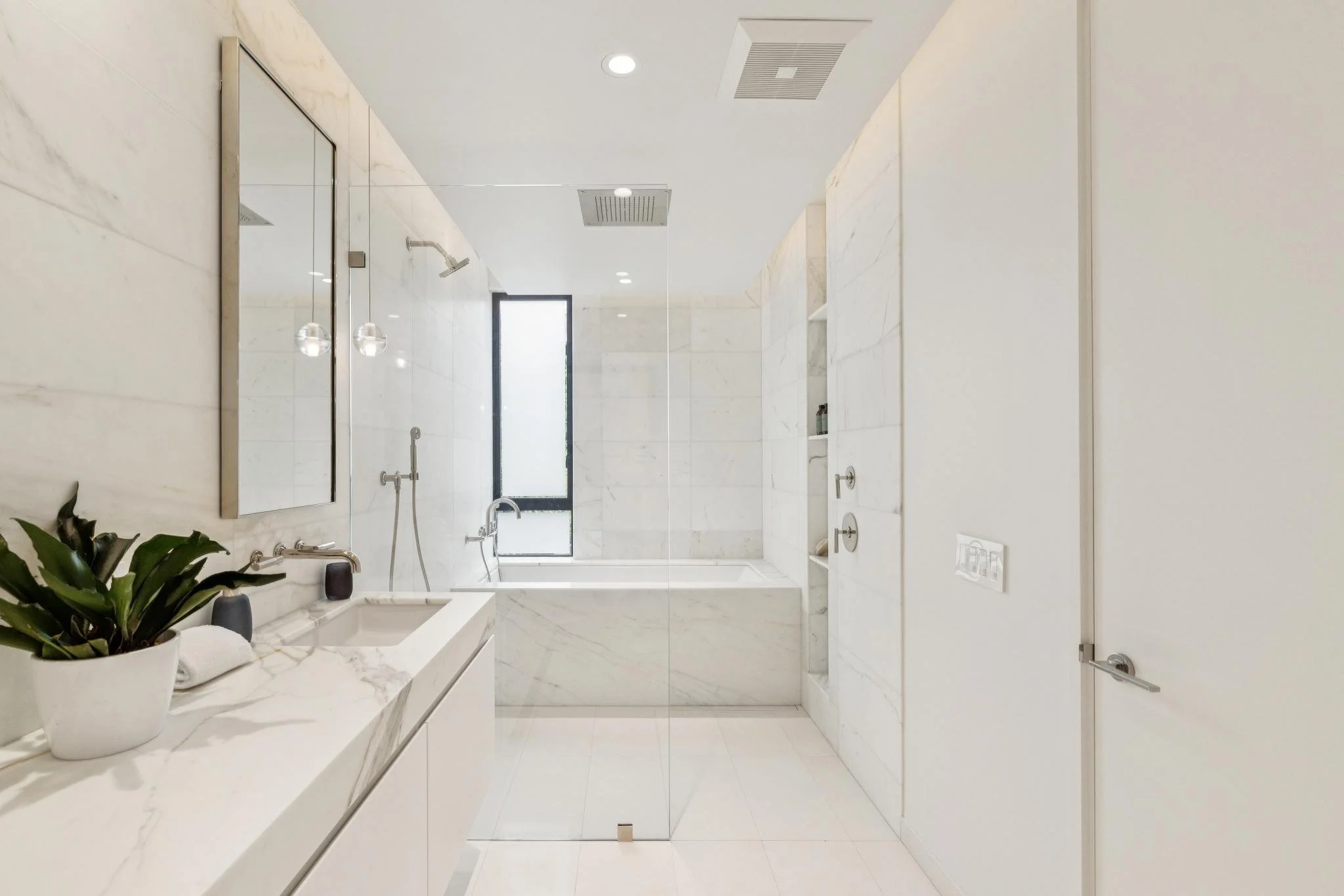 Modern bathroom with white marble walls and floors, a large mirror above the sink, a potted plant, and a walk-in shower with glass walls and a window, ceiling lights, and a towel bar.