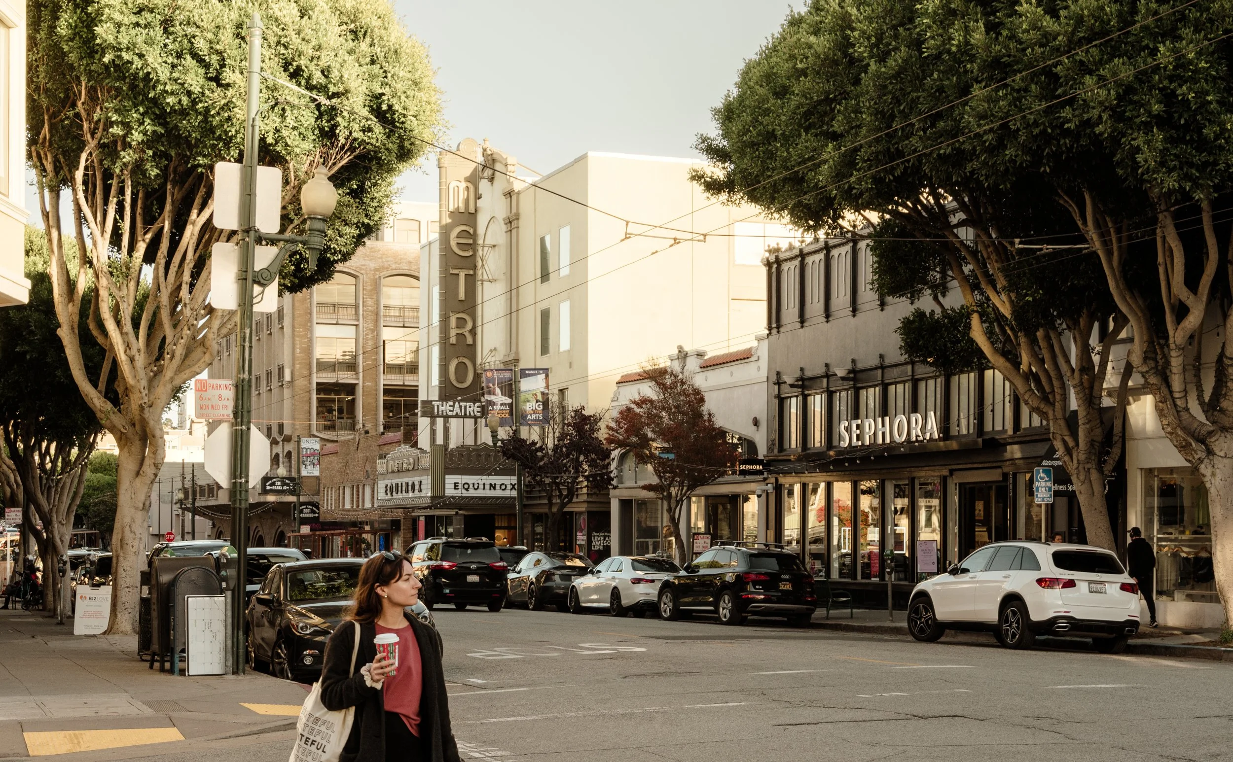 Street scene with cars parked along the curb, trees with lush green foliage, and storefronts including Sephora and Equinox; a woman holding a drink with a patterned tote bag walking across the street.