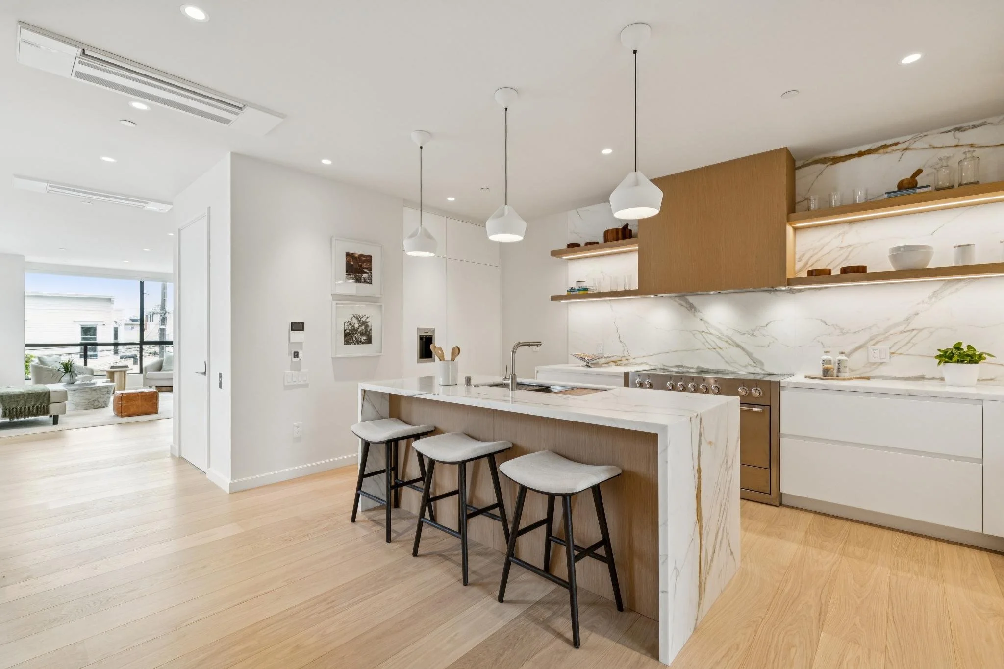 Modern open-concept kitchen with marble island, pendant lighting, and light wood cabinets, opening to a living area with large windows.