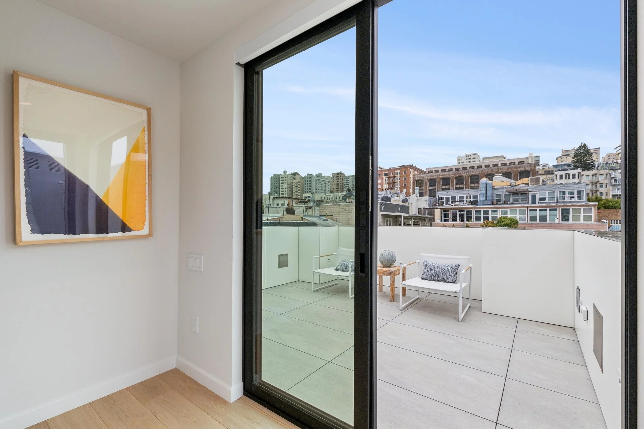 A modern apartment interior with a sliding glass door leading to a balcony. The balcony has two white chairs with patterned pillows and a small wooden table with a spherical decor piece. The city skyline can be seen through the window with multiple b