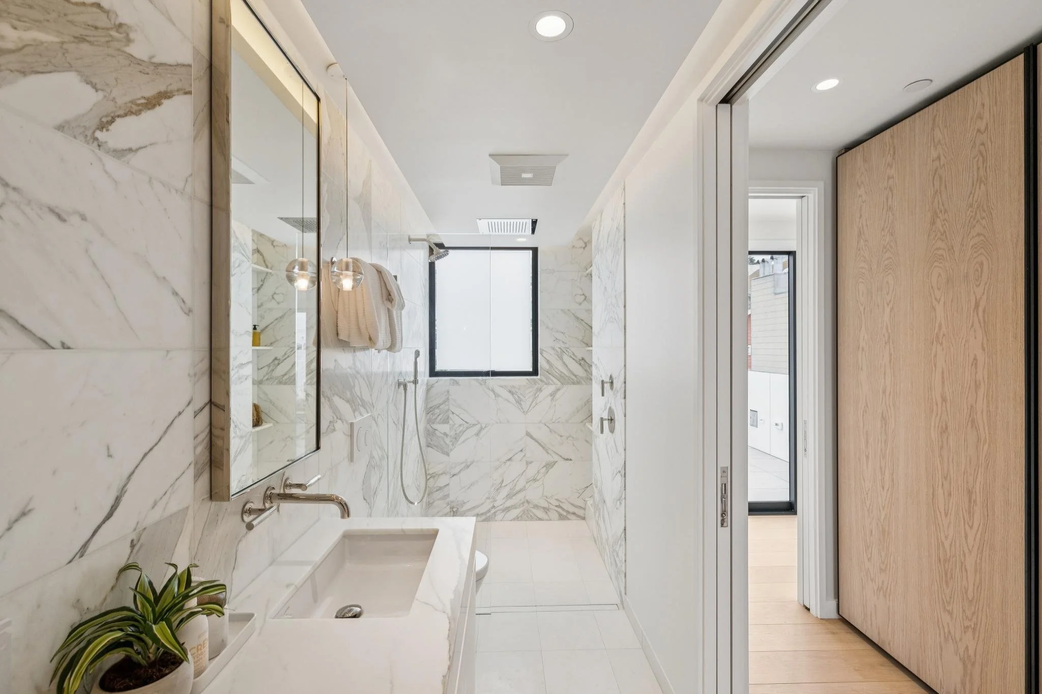 A modern bathroom with marble walls and floor, a double sink vanity with a mirror, two pendant lights, a window at the back, and an open door leading to another room.