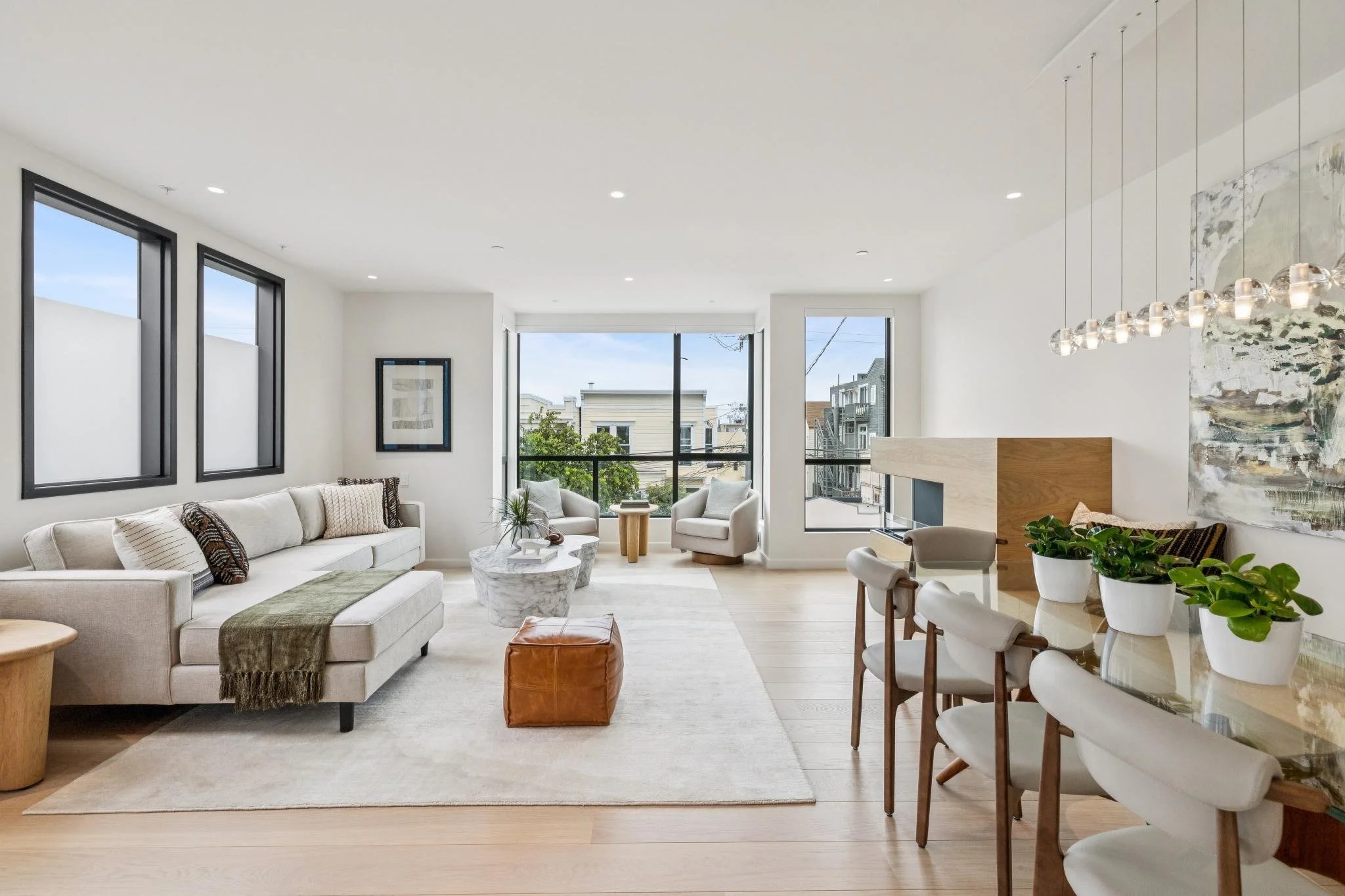 Bright living room with large windows, beige sofa with patterned cushions, armchairs, marble coffee table, plants, modern chandelier, and a view of neighboring houses outside.