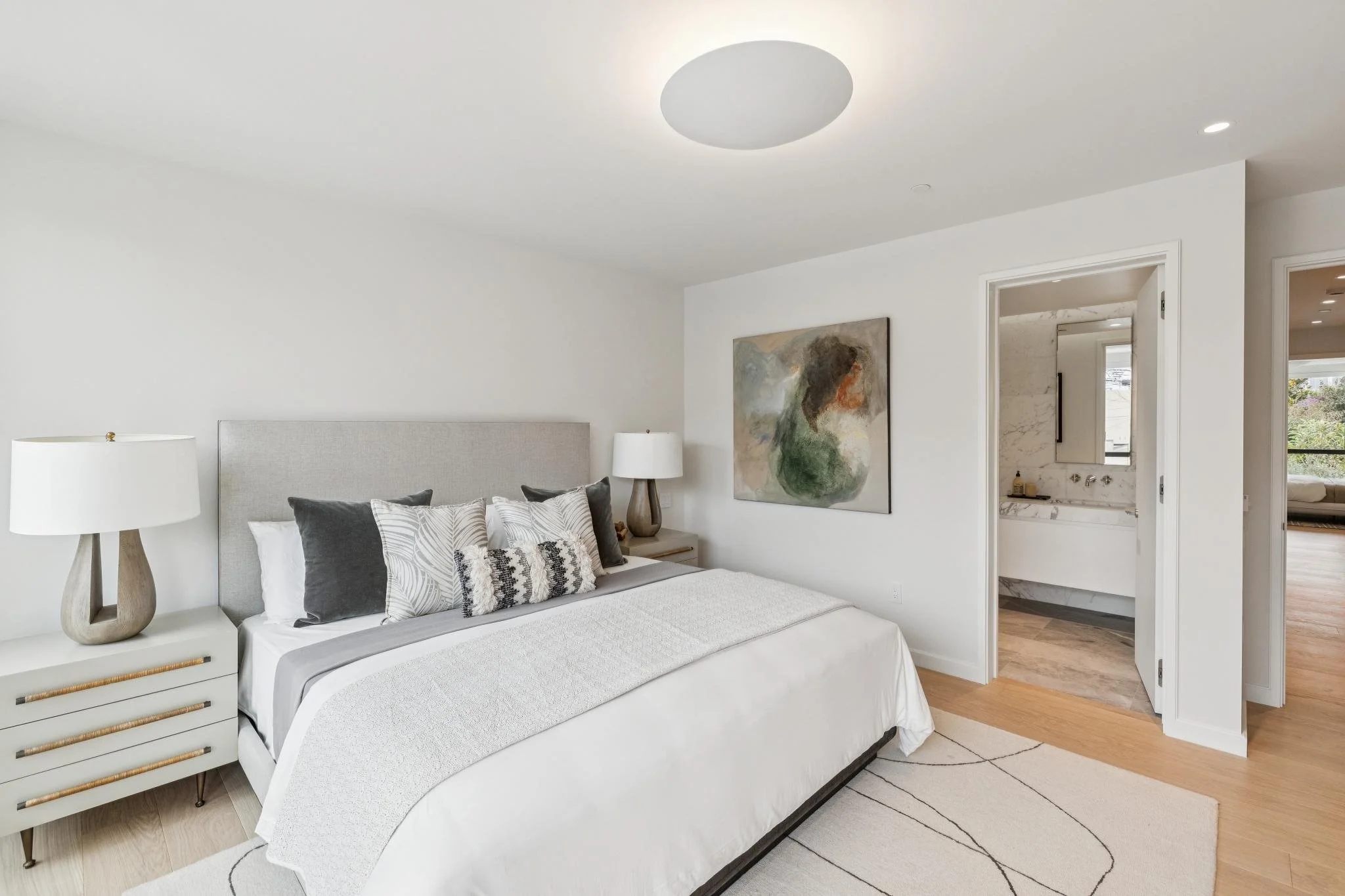 Modern bedroom with a bed, nightstand, table lamp, wall art, and an adjacent bathroom.
