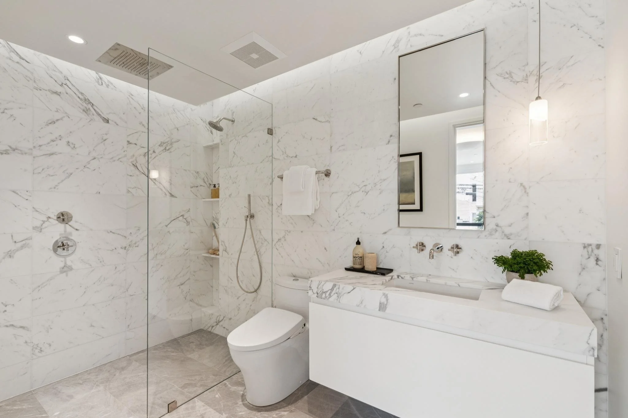 Bathroom with marble walls and floor, shower area with glass partition, white toilet, marble vanity with soap bottles, white towel, wall mirror, a window, and a hanging pendant light.