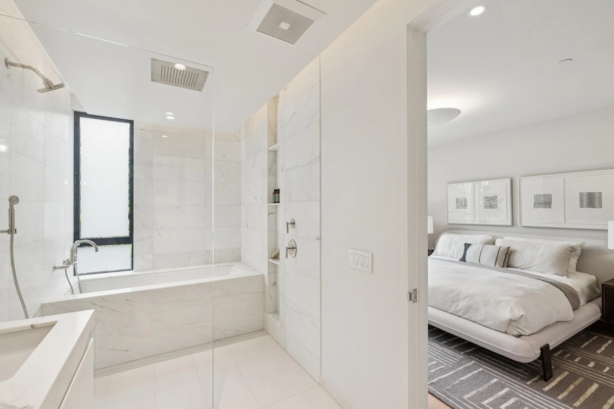 Modern bathroom with white marble tiles and large window, adjacent to bedroom with white bed, pillows, and framed artwork on wall.