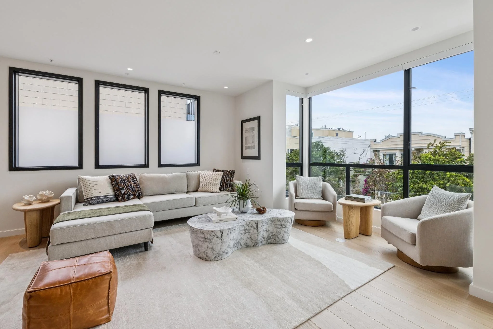 Bright living room with large windows, white walls, cream sofa with patterned pillows, white armchair, marble coffee table, wooden side tables, and greenery outside.