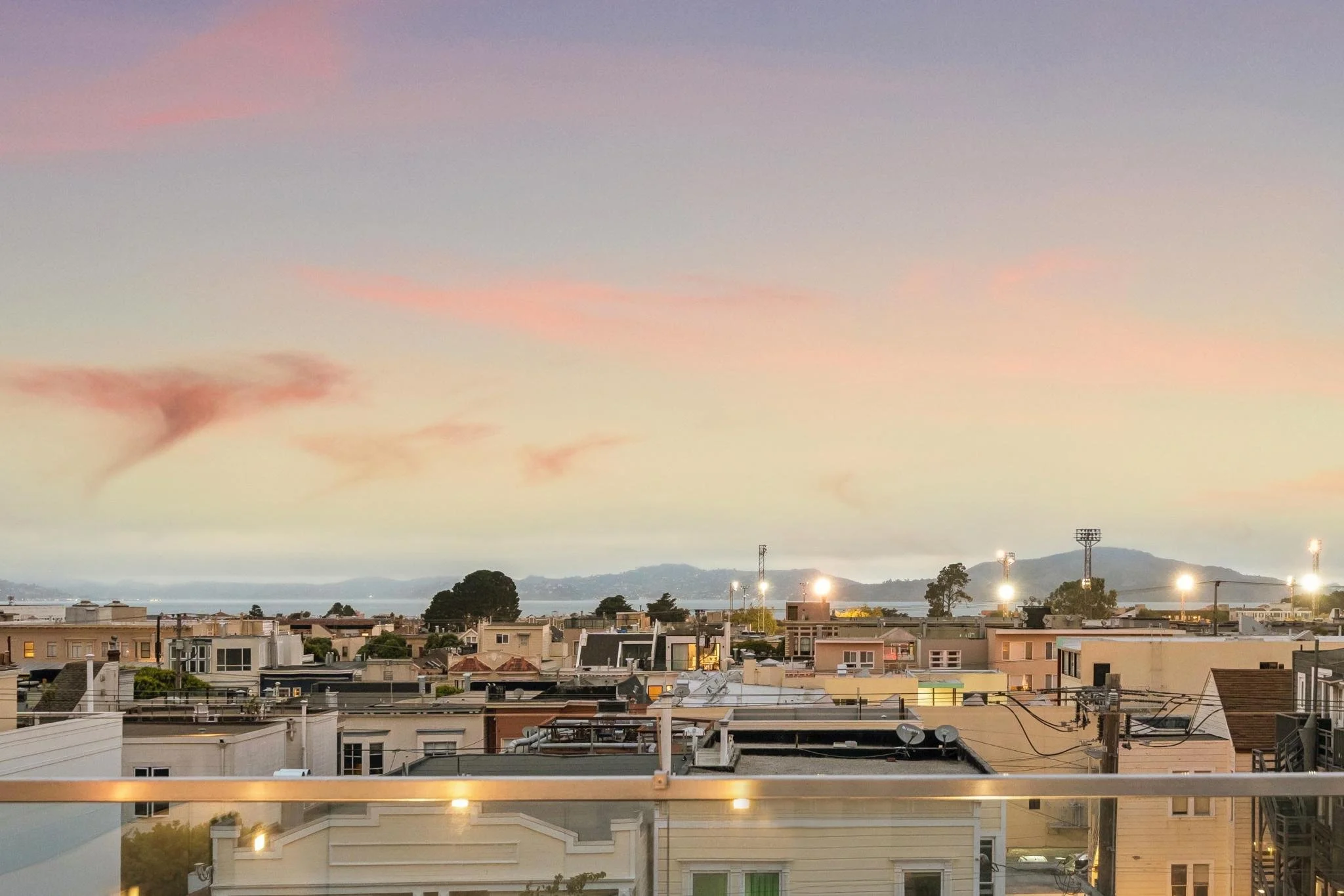 Cityscape view during sunset with residential rooftops, trees, and hills in the background, under a sky with pink and purple clouds.