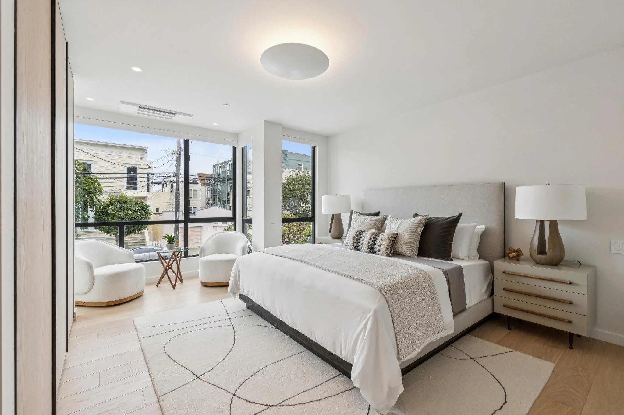 Bright bedroom with large windows showing an urban neighborhood, featuring a bed with neutral-colored pillows, two nightstands with lamps, and a white area rug with black curved lines.