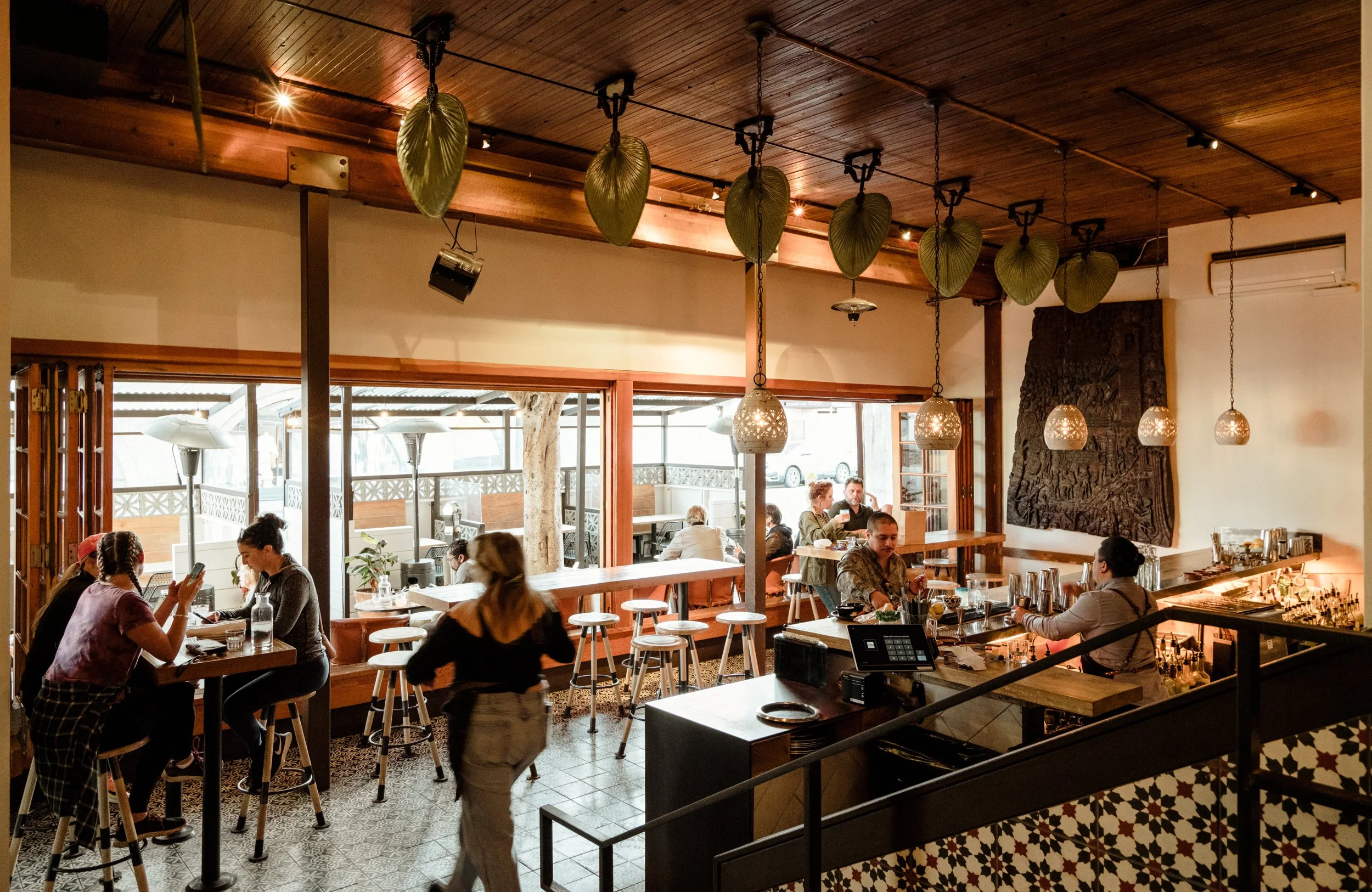 Interior of a cozy restaurant with high wooden ceiling, hanging pendant lights, and large windows. Several groups of people are seated at tables enjoying meals, with some engaged in conversation and others using their phones. The decor includes green