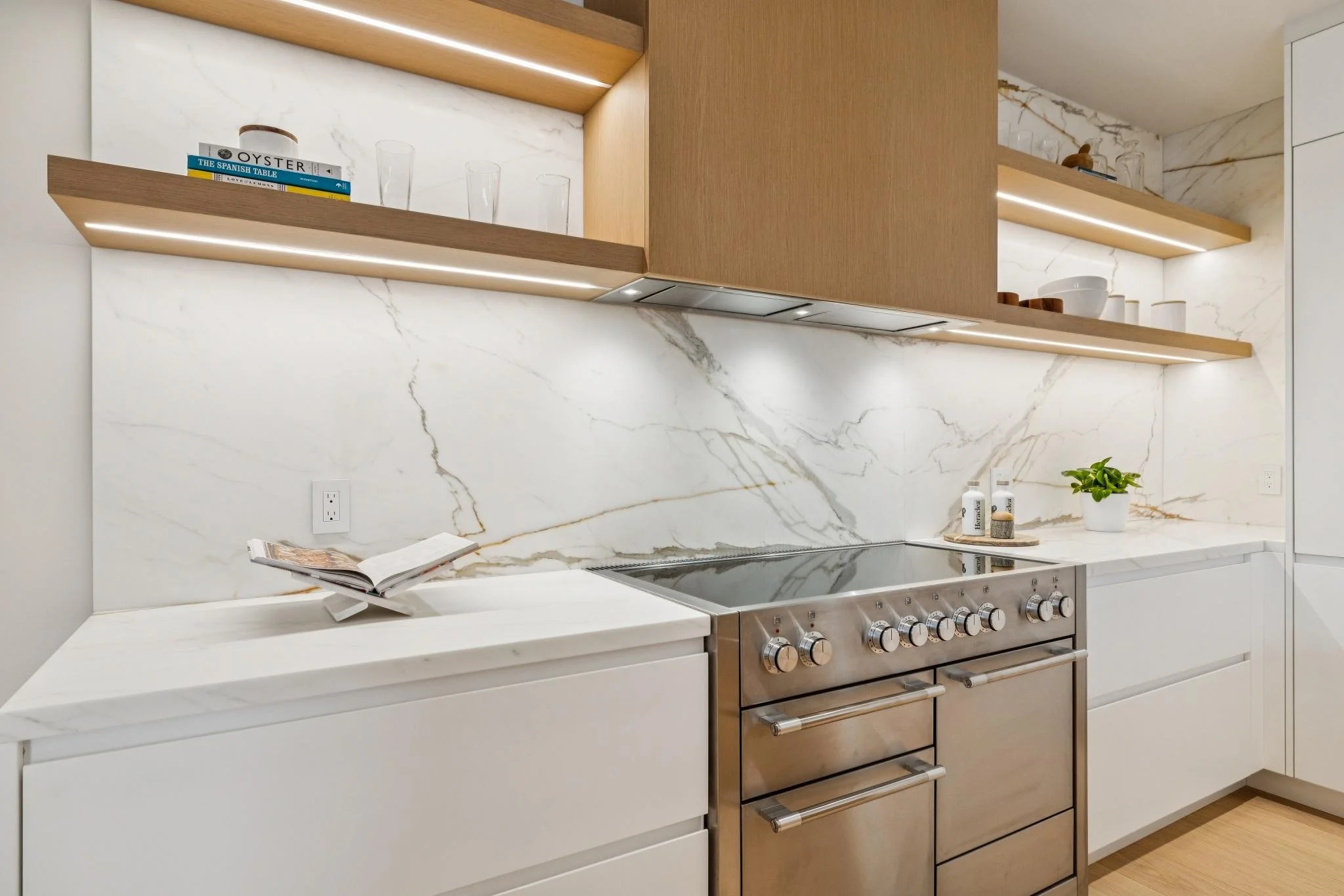 Modern kitchen with white marble backsplash, wooden open shelves, stainless steel stove, and minimal decor including glassware, bowls, a plant, and books.