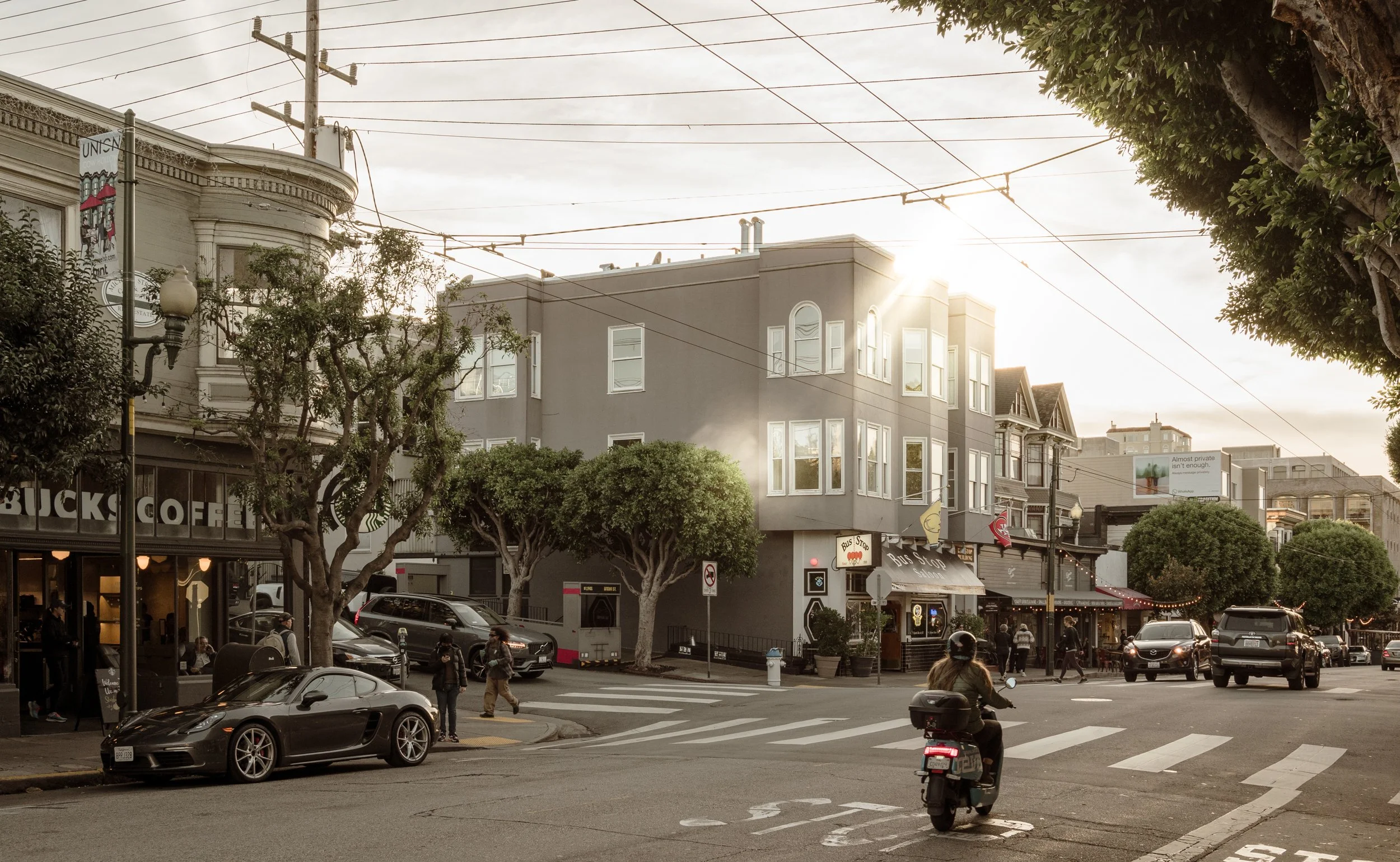 City street scene with cars, pedestrians, trees, and multi-story buildings, including a Starbucks coffee shop, with overhead wires and a cloudy sky sunlight.