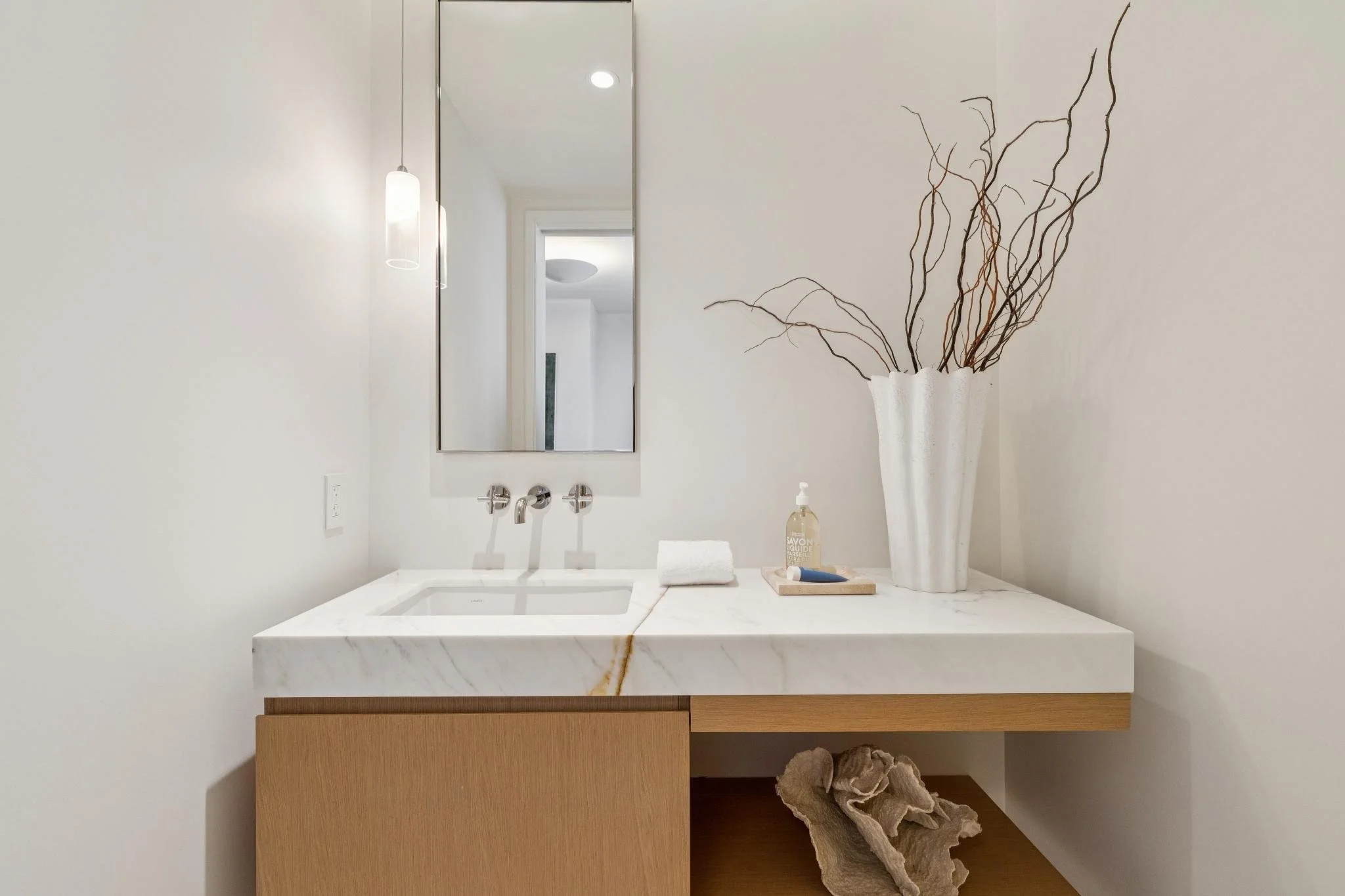 A modern bathroom vanity with a white marble countertop, a rectangular sink, and a large mirror. The vanity has a wooden cabinet below, decorated with a white towel, a bottle of liquid soap, and a tray. To the right, a tall white vase with decorative