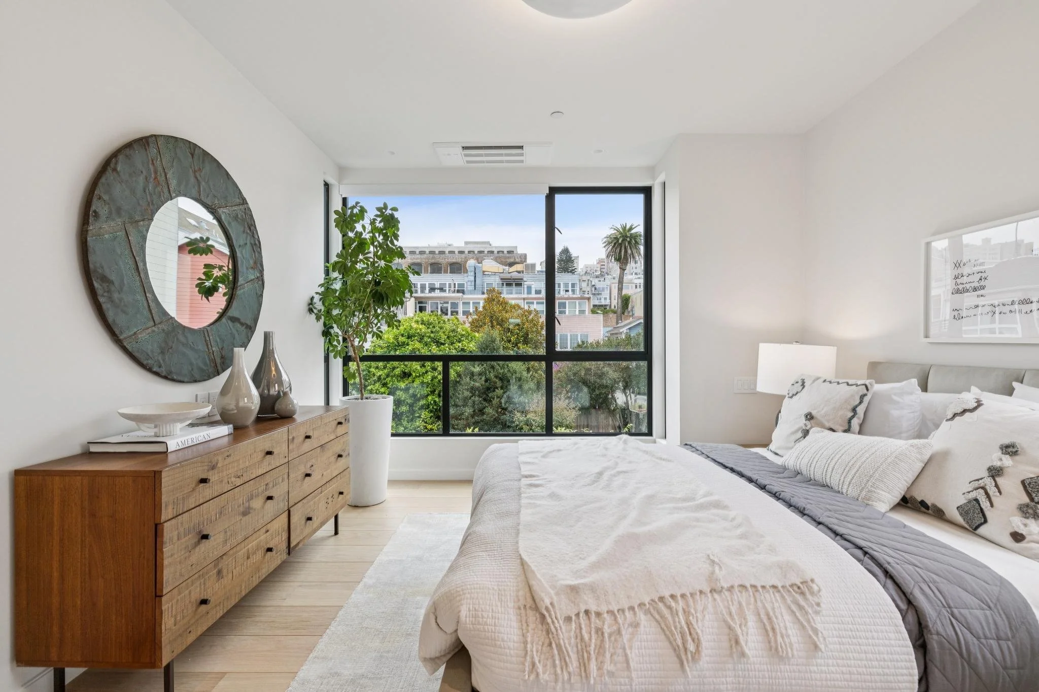 A bright bedroom with a large window showing a cityscape and trees outside. There is a bed with white and gray bedding, a wooden dresser with decorative vases, a mirror, and a potted plant near the window.