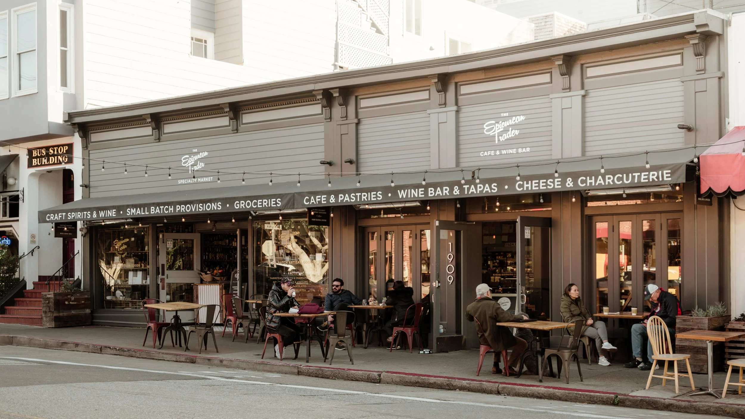 Exterior view of a cafe and wine bar named The Epicurean Trader, with outdoor seating and people dining outside. The signage indicates offerings like craft spirits, wine, groceries, pastries, cheese, and charcuterie.