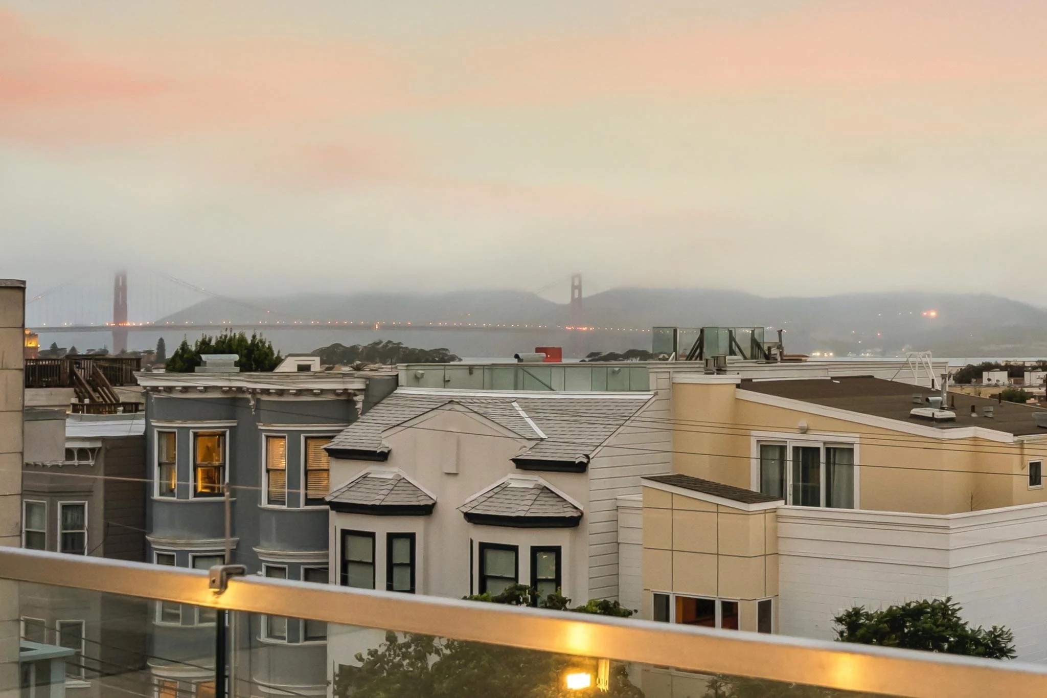 View of rooftops of residential buildings with a bridge in the background over water, possibly the Golden Gate Bridge, during dusk or dawn with soft pink and blue sky.