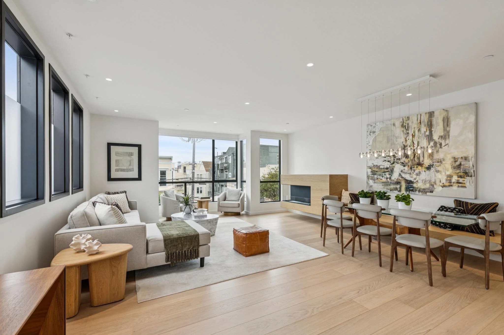 Open living and dining area with modern decor, large windows, artwork, beige sofas, wooden furniture, and potted plants.