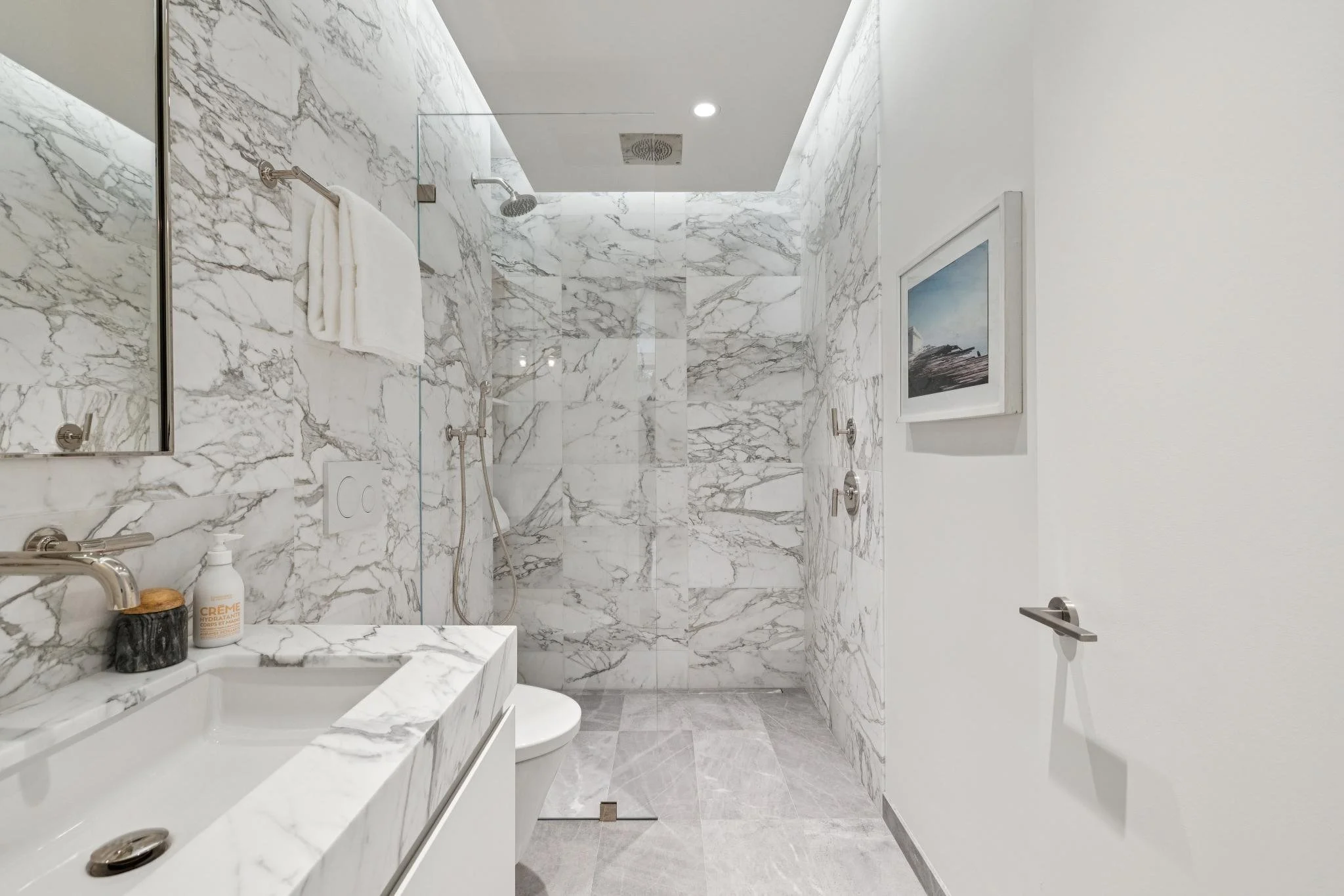 A modern bathroom with marble walls and floor, a walk-in shower with a glass door, a bathtub, a towel hanging on a wall-mounted towel bar, a framed picture on the white wall, and a faucet with toiletries on a marble vanity.