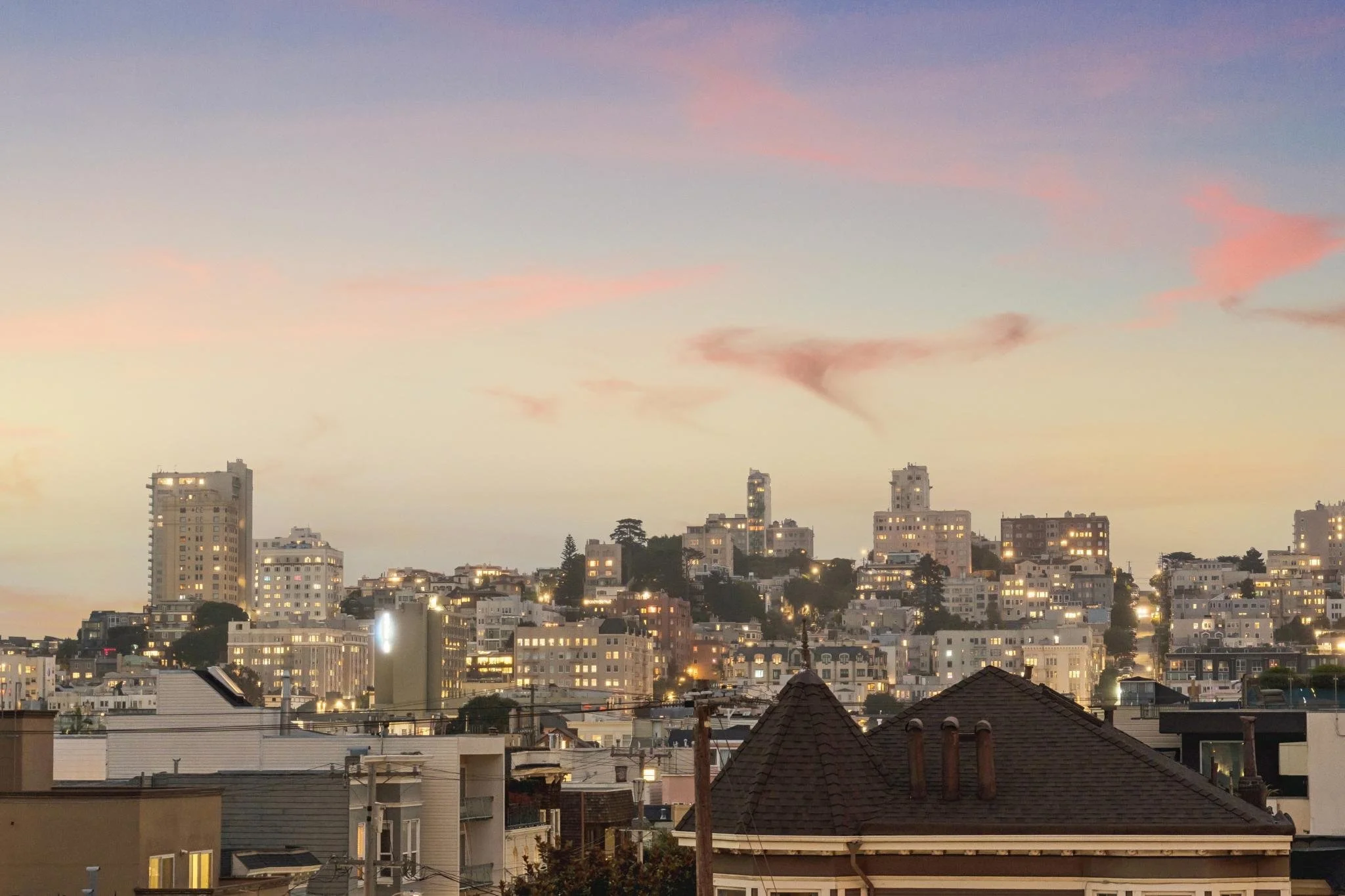 A cityscape at dusk with high-rise buildings, some lit windows, and hills with houses and trees in the background. The sky has pastel-colored clouds.