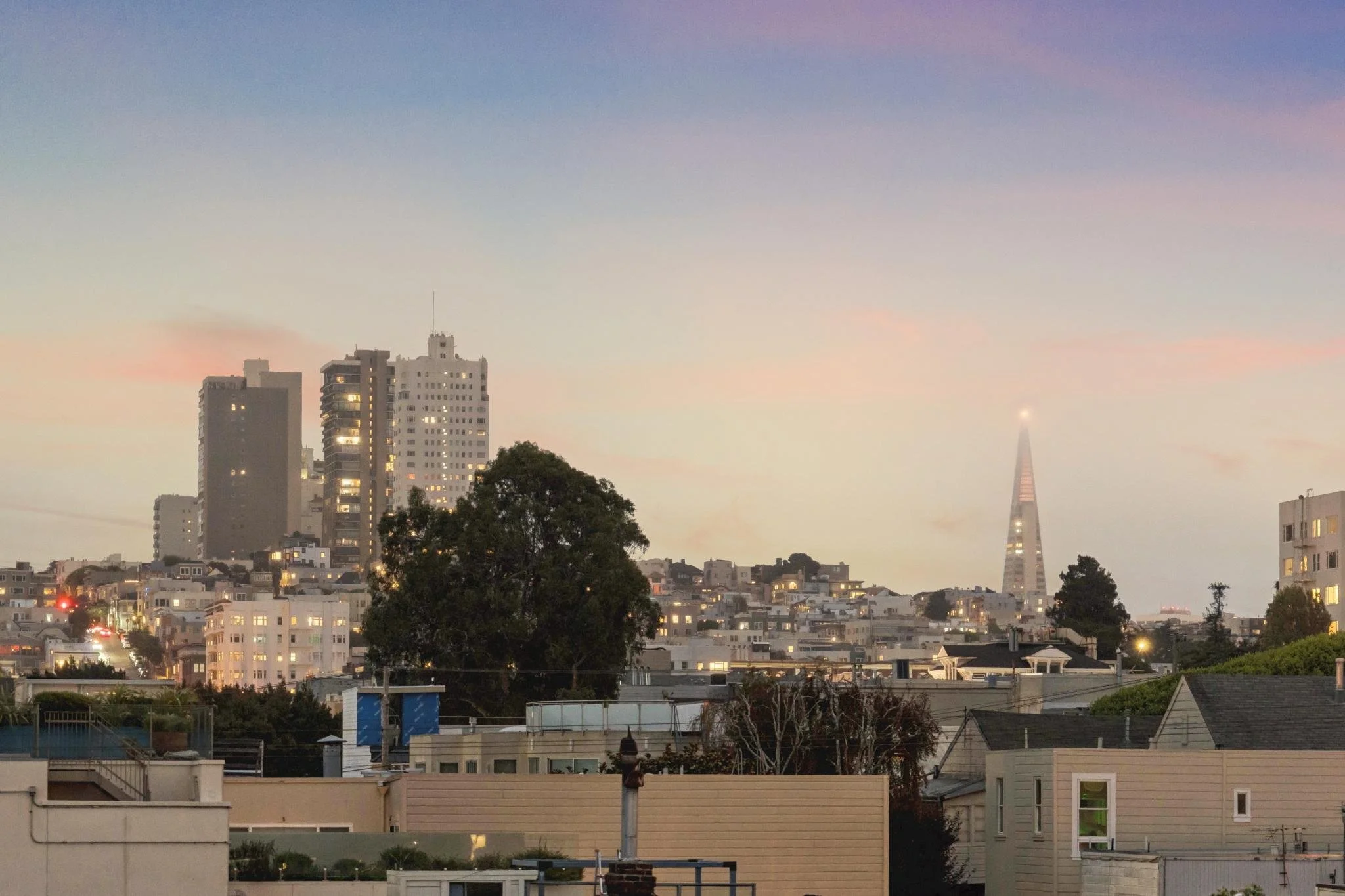 Cityscape at dusk with buildings, trees, and the Salesforce Tower in San Francisco in the background under a colorful sky.