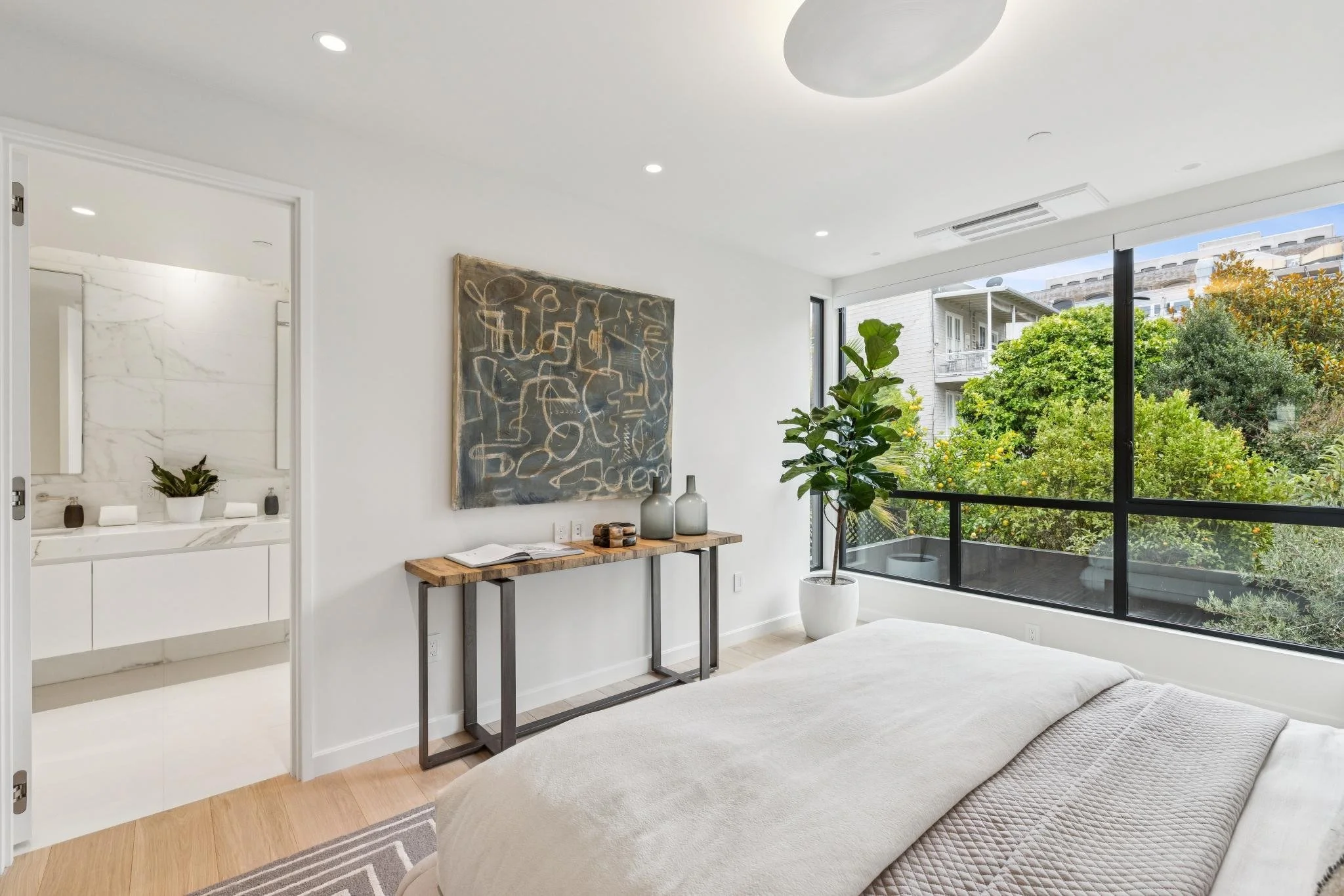 Contemporary living room with large window overlooking greenery, minimalistic artwork on white wall, potted plant, and natural wooden console table with decorative items.