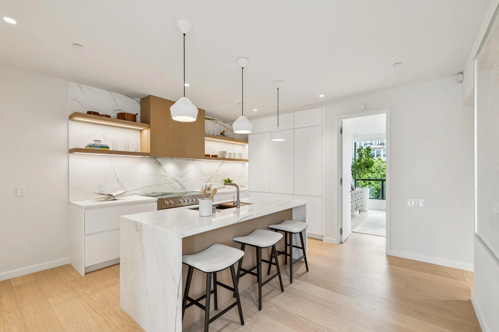 Modern kitchen with white marble island and backsplash, wooden open shelves, black bar stools with white cushions, and large window with greenery outside.