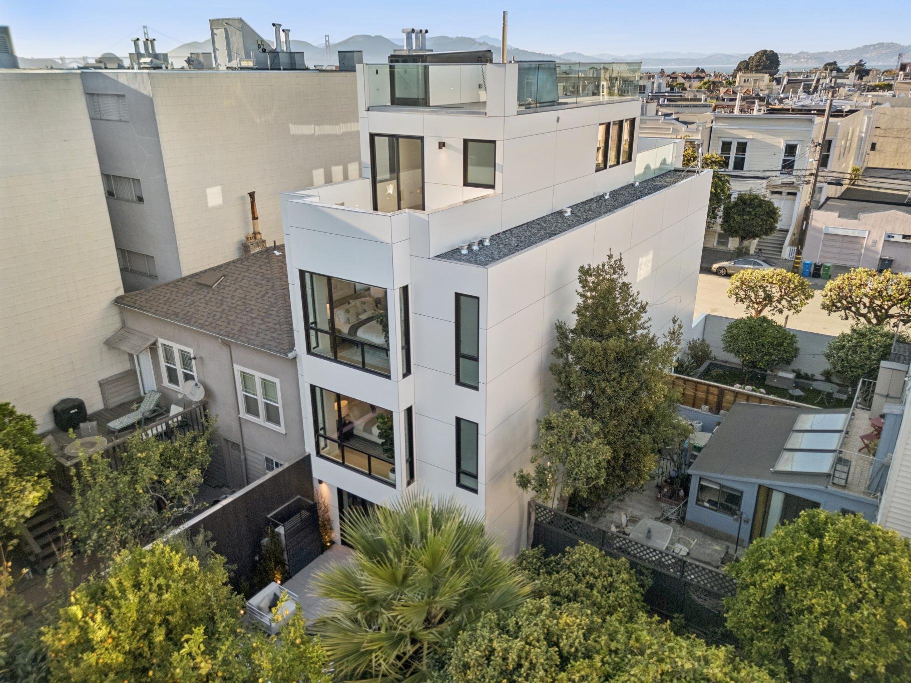 A modern multi-story white apartment building with glass balconies located in a densely built urban neighborhood, surrounded by trees, older houses, and city streets with cars, in San Francisco with distant hills in the background.