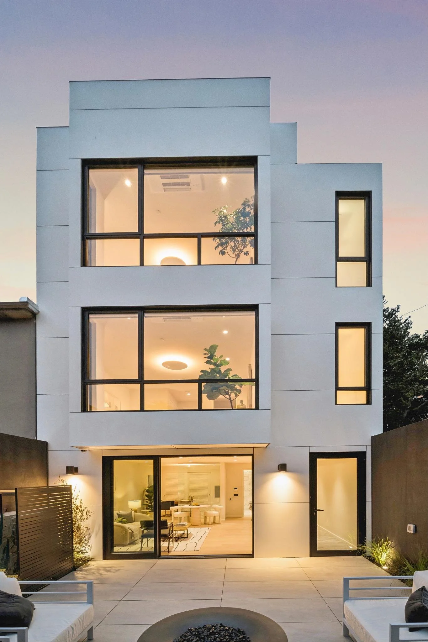 Modern white residential building with large windows illuminated from inside, outdoor patio area with white sofas and a round fire pit, evening sky backdrop.