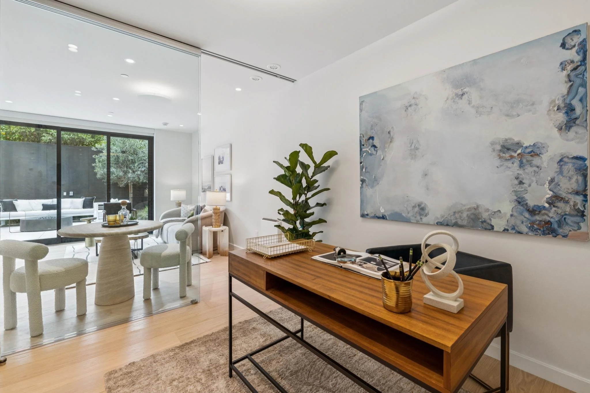 Modern living room with white walls, wooden floor, and large sliding glass doors leading to an outdoor patio with white outdoor furniture. Interior includes a round table with four upholstered chairs, a wooden sideboard with decorative items, and a l