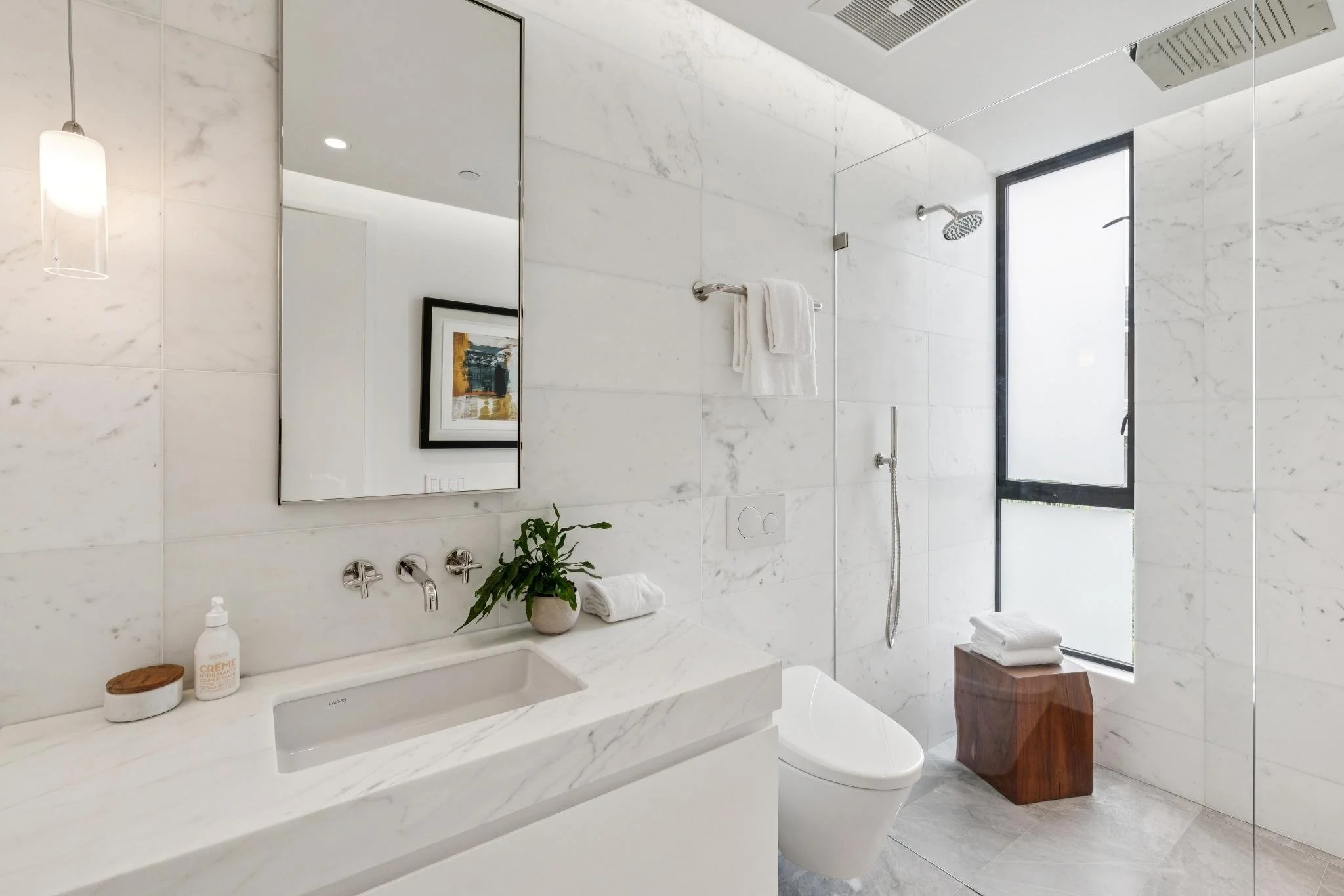 A modern bathroom with white marble walls and countertops, a rectangular mirror, a potted plant, a bottle of cream, folded towels, a toilet, and a walk-in shower with a black-framed window and a rainfall showerhead.