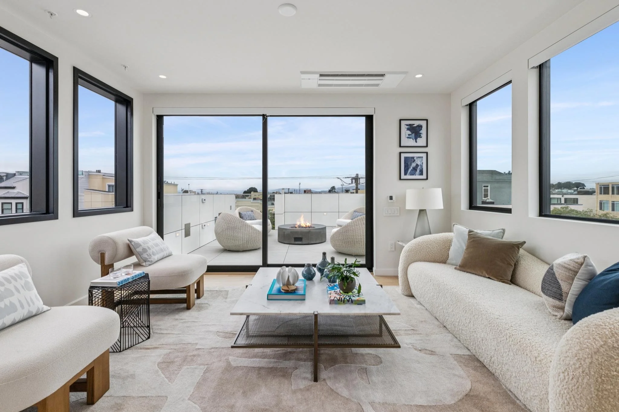 Modern living room with large windows, a white sofa, chairs around a coffee table, and a balcony with outdoor seating and a fire pit, cityscape view