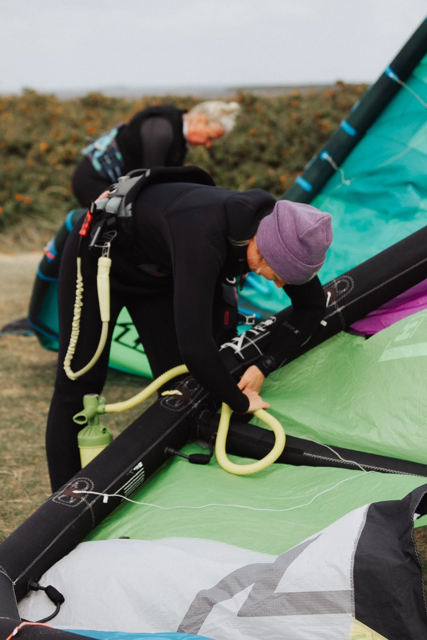 Zwei Menschen bereiten gemeinsam ein Kitesurfsegel vor. Eine Person ist gebückt und arbeitet an der Verbindung des Segels, die andere Person im Hintergrund steht an einem Strand.