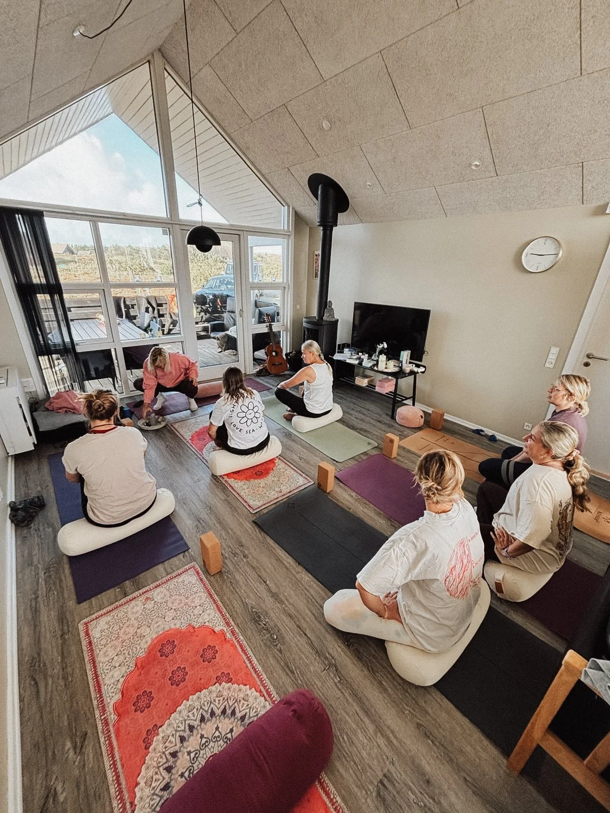 Menschen machen eine Yogastunde in einem hellen Raum mit großen Fenstern, Sofa, Fernseher und Musikinstrumenten.