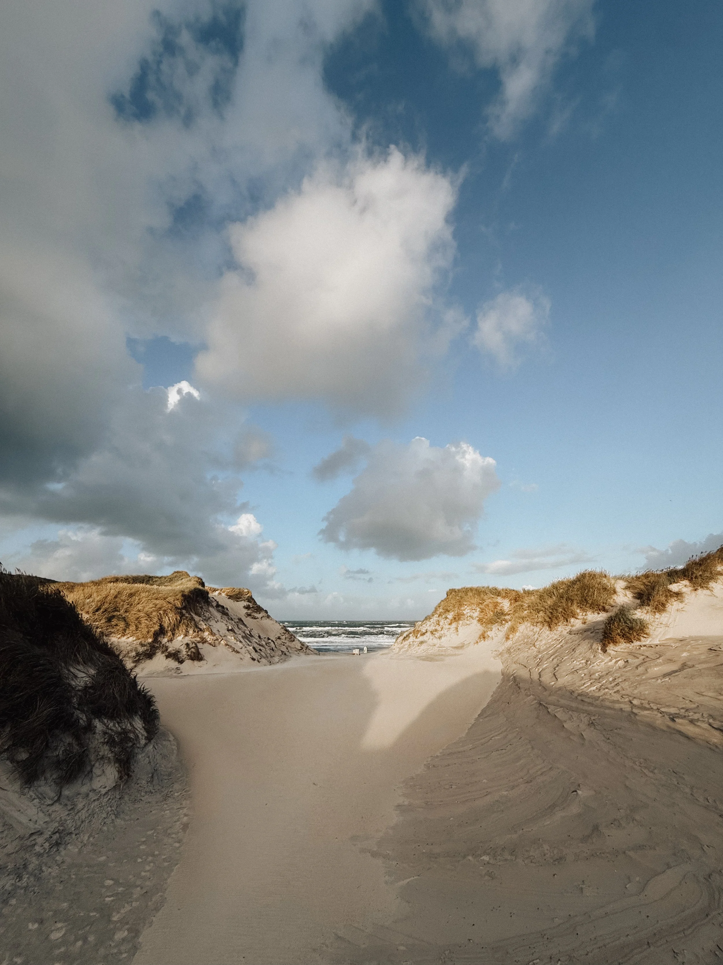Ein Sandstrand zwischen Dünen mit Grasbewuchs und das Meer im Hintergrund, unter einem bewölkten Himmel.