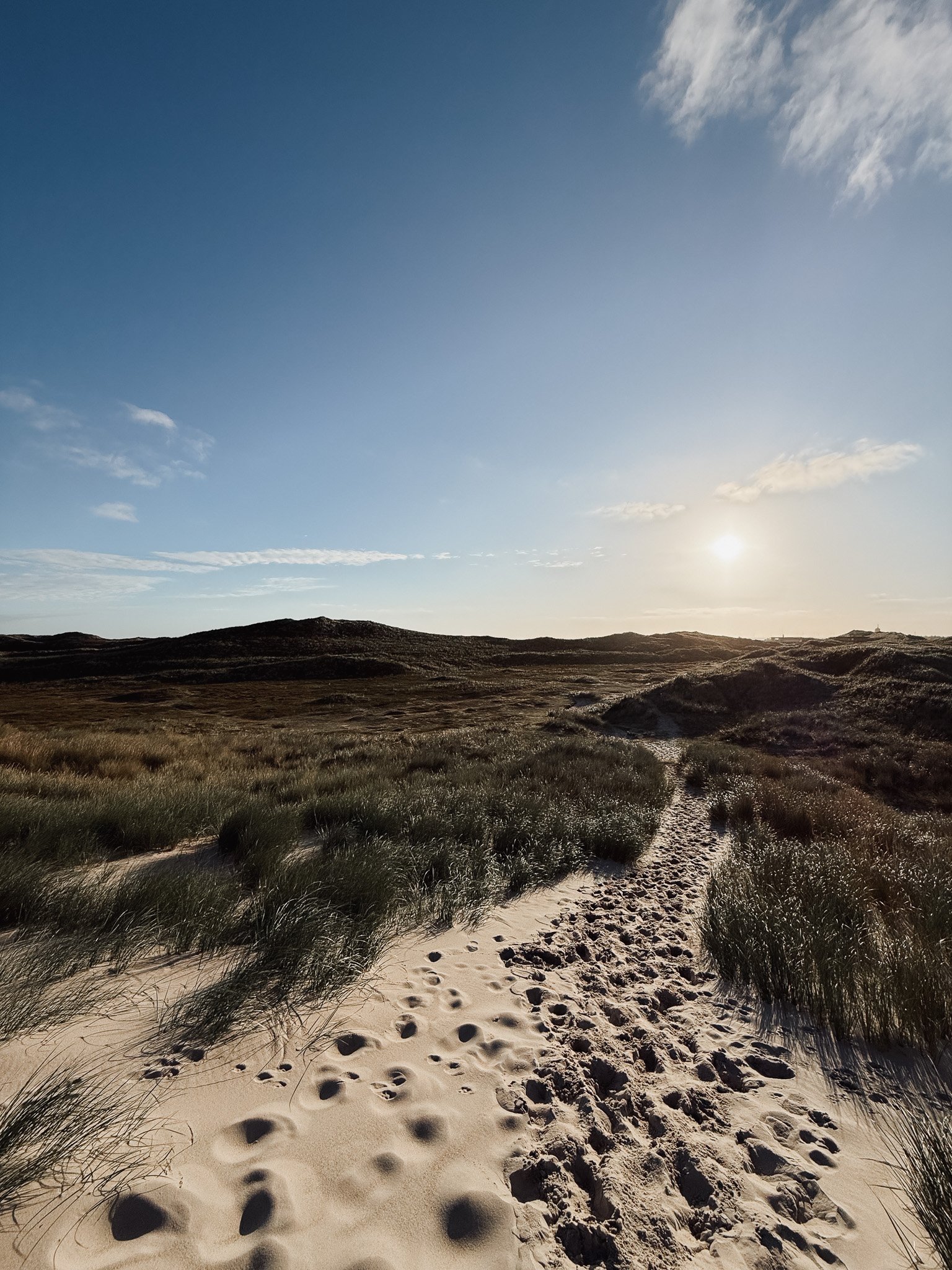Ein Sandpfad führt durch Dünen mit Gras, bei Sonnenuntergang oder -aufgang, hinter einem Hügel mit einem kleinen Gebäude in der Ferne, Himmel mit einigen Wolken.