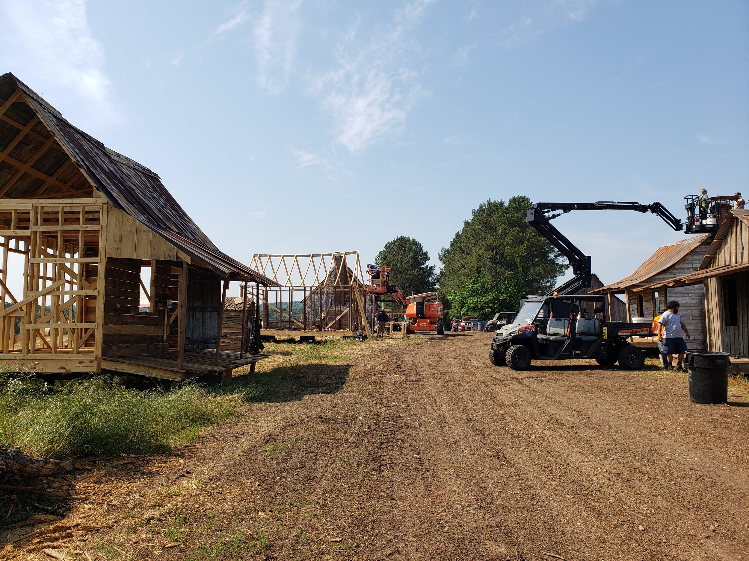 Row of period cabins under construction on a farm film set, some still bare framing, with a boom lift and utility vehicle on the dirt lane