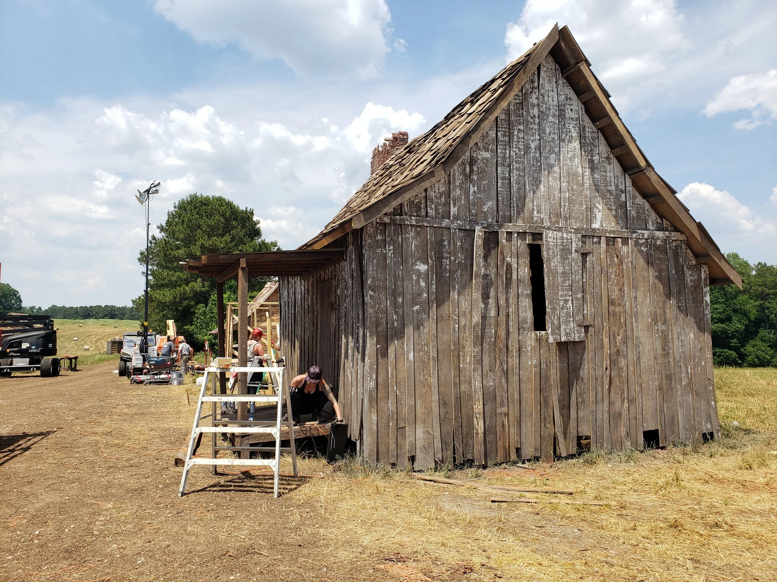Weathered gray wood cabin with a shake roof and stone chimney on a period farm film set, a crew member working at its base beside a stepladder