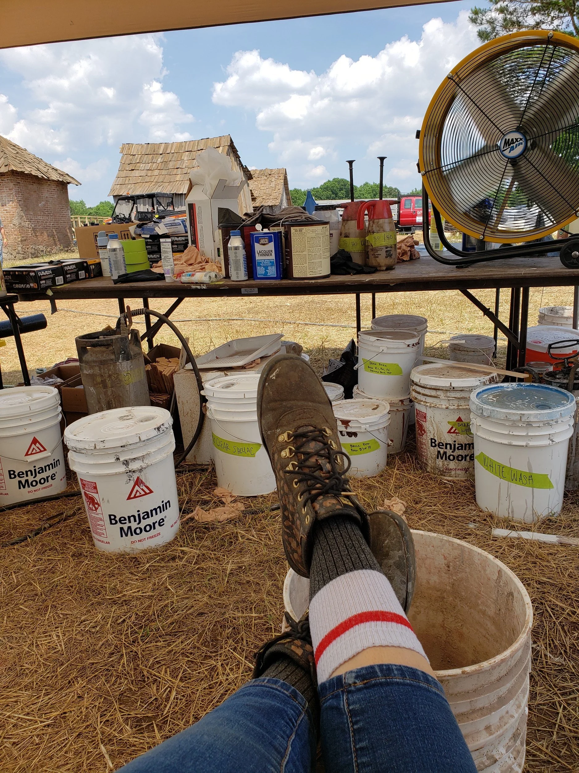 Paint station under a shade tent on a period farm film set: labeled paint buckets, a supply table and work boots propped on a bucket, with cabins behind