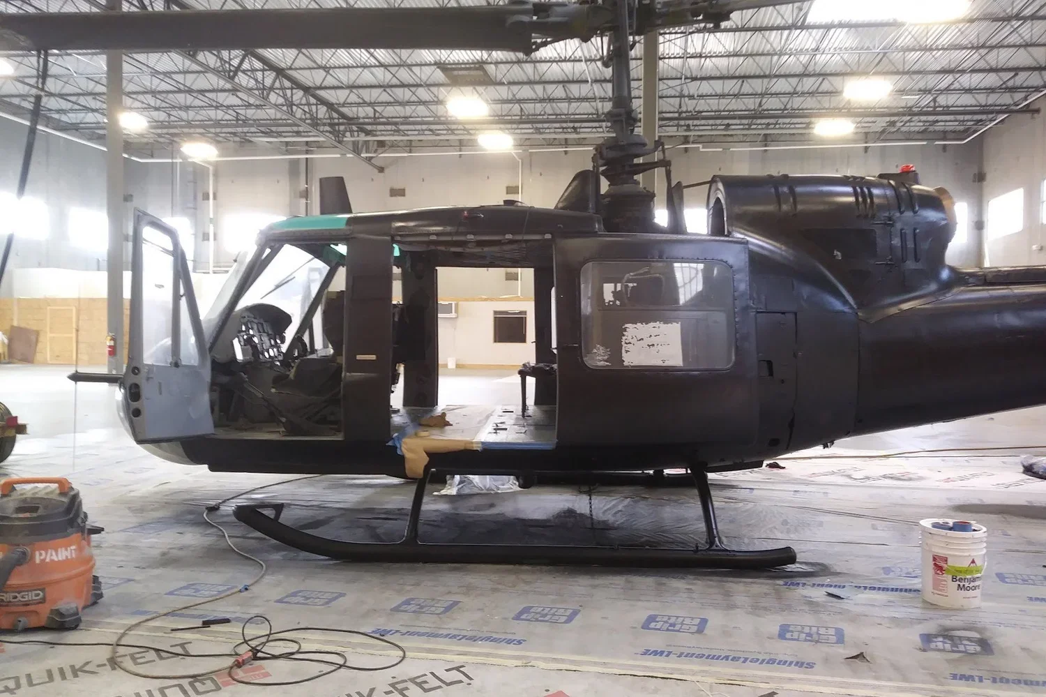 Full-size helicopter sprayed black inside a studio, doors open, on a paper-covered floor with a shop vac and paint bucket nearby