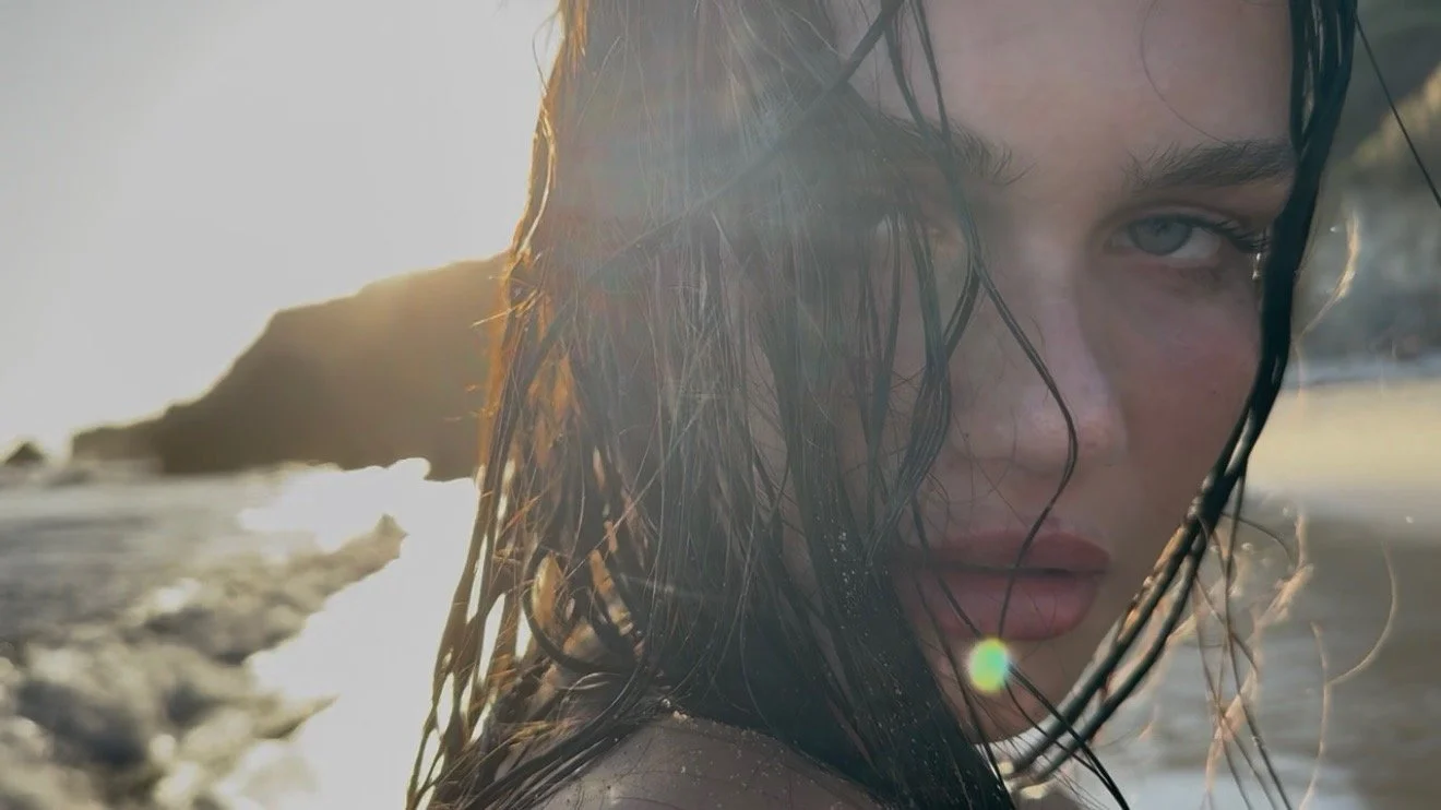 Close-up of a woman with wet hair and blue eyes by the beach at sunset, looking into the camera.
