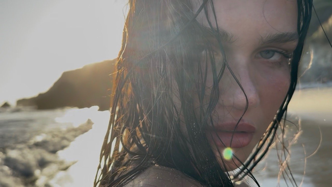Close-up of a woman with wet hair at the beach during sunset, looking into the camera.