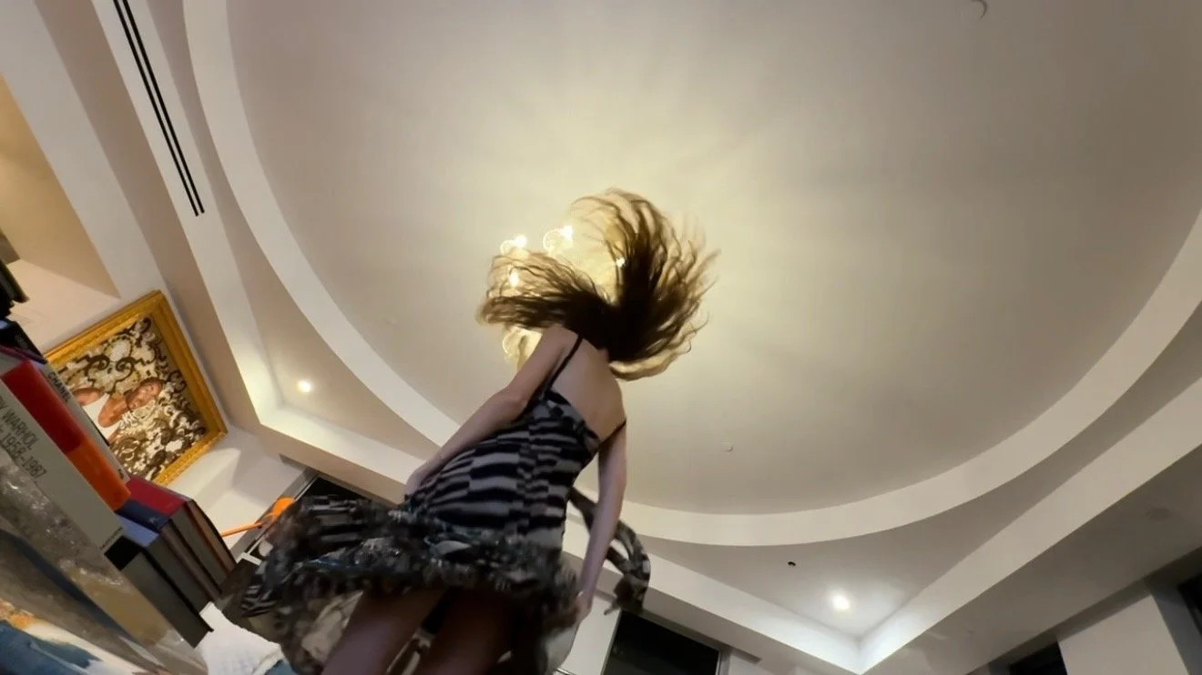 A woman with long hair wearing a black and white striped dress, caught mid-spin, inside a room with a curved ceiling, a framed picture on the wall, and a table with books and a can of beer.