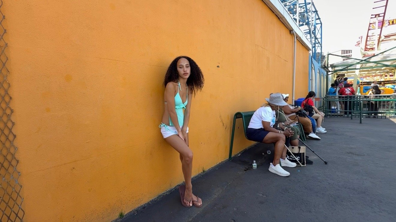 A young woman with curly hair wearing a light blue top and shorts stands against an orange wall at an amusement park. Several older women are sitting on benches nearby, some wearing hats and sunglasses, with one holding a cane. In the background, amu