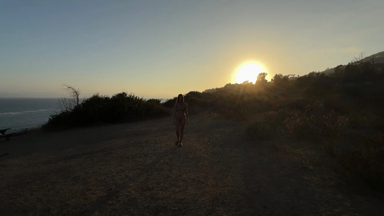 A woman walking along a dirt path near the coast during sunset, with the sun low over the horizon and bushes on either side.