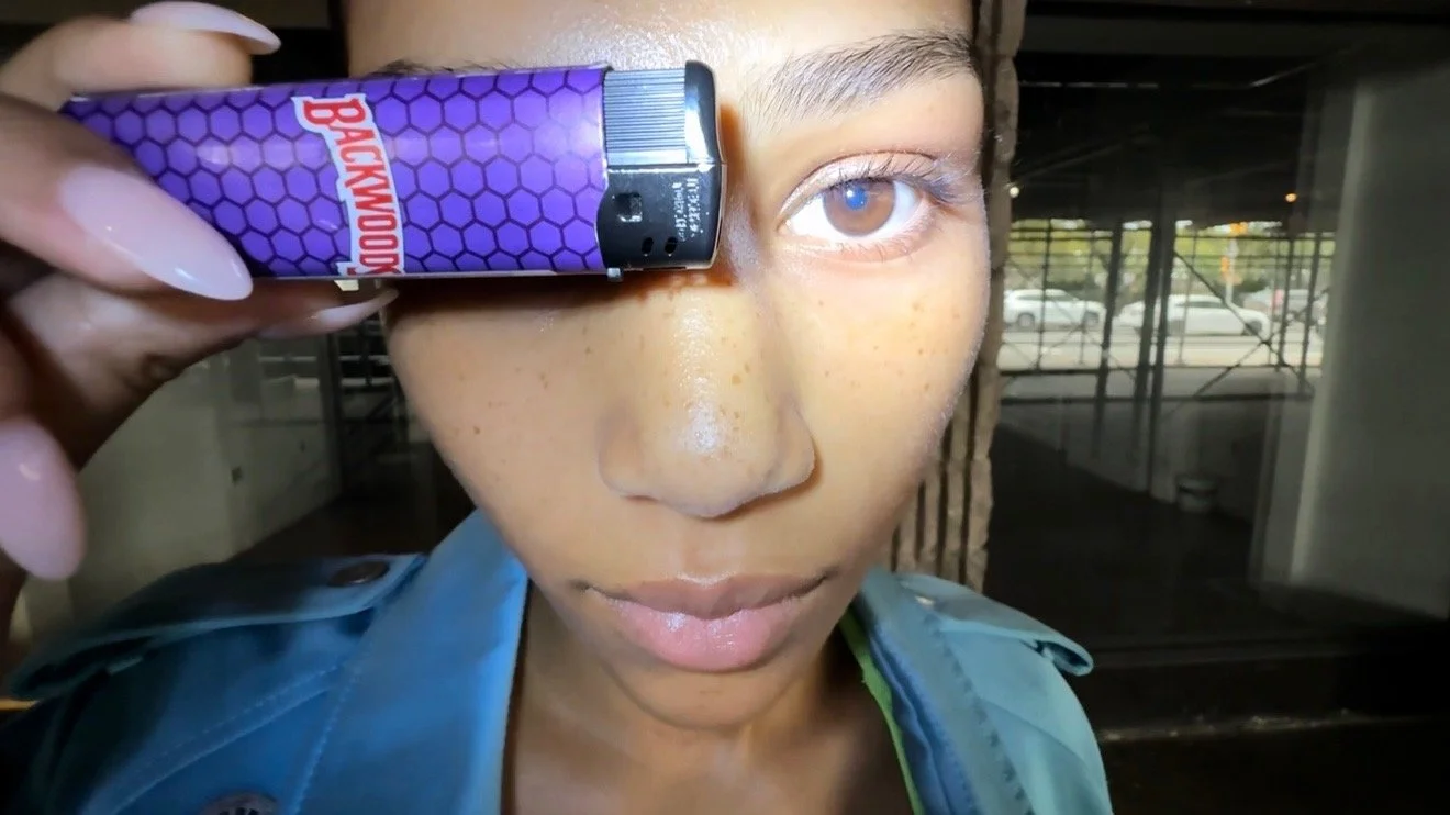 Person holding a purple Backwood cigarette near their left eye inside a building with large windows.