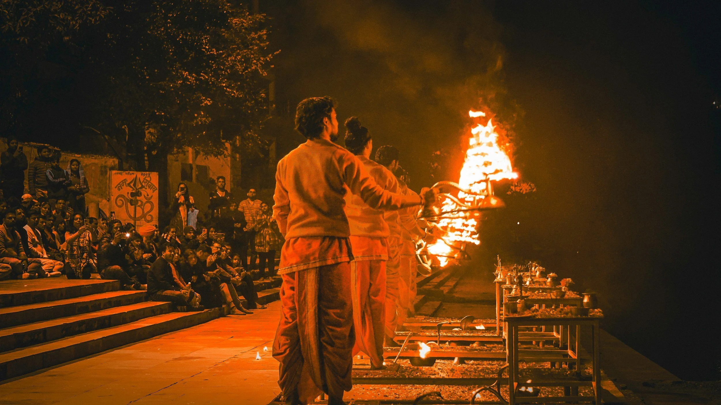 Group of people participating in a traditional fire ritual at night, with a large bonfire and onlookers sitting on steps.