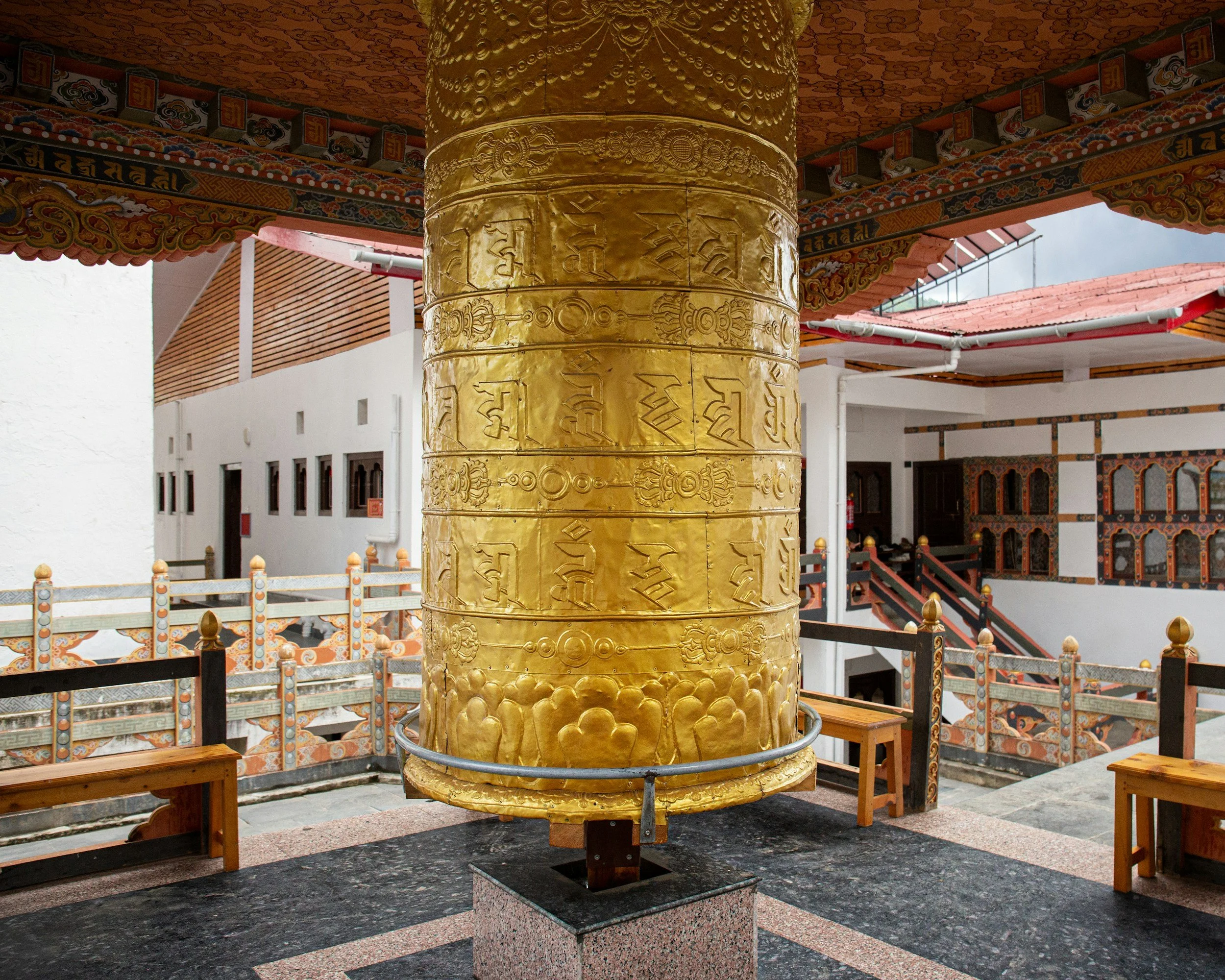 A large golden prayer wheel in the center of a temple, decorated with intricate patterns and inscriptions, surrounded by wooden benches and ornate architectural details.